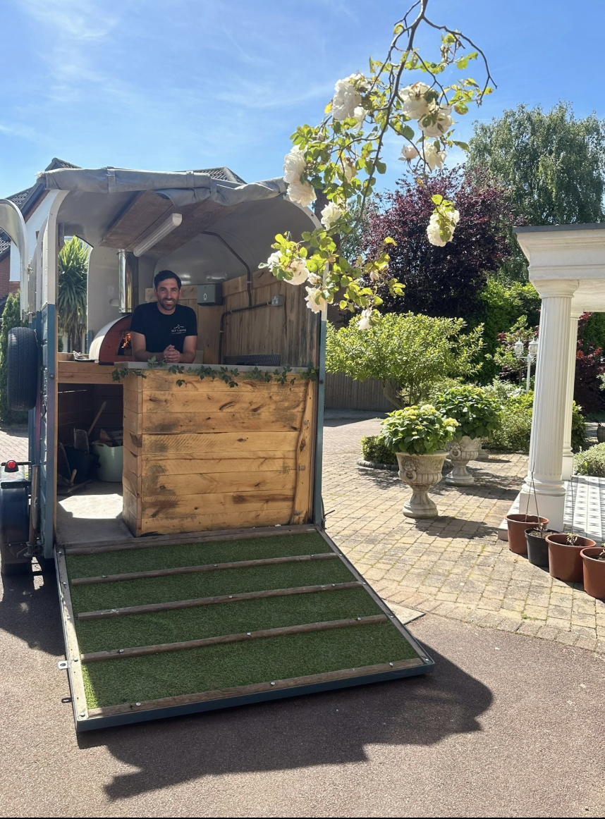 A vintage catering van features a wooden serving counter, lush greenery, and a smiling attendant in bright daylight, enhancing the inviting atmosphere.