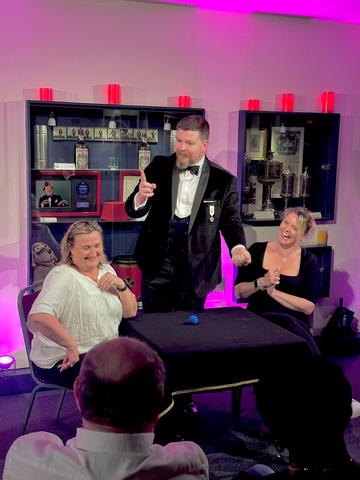 The magician, dressed in a tuxedo, performs on a small stage with dramatic purple lighting, engaging two amused audience members.