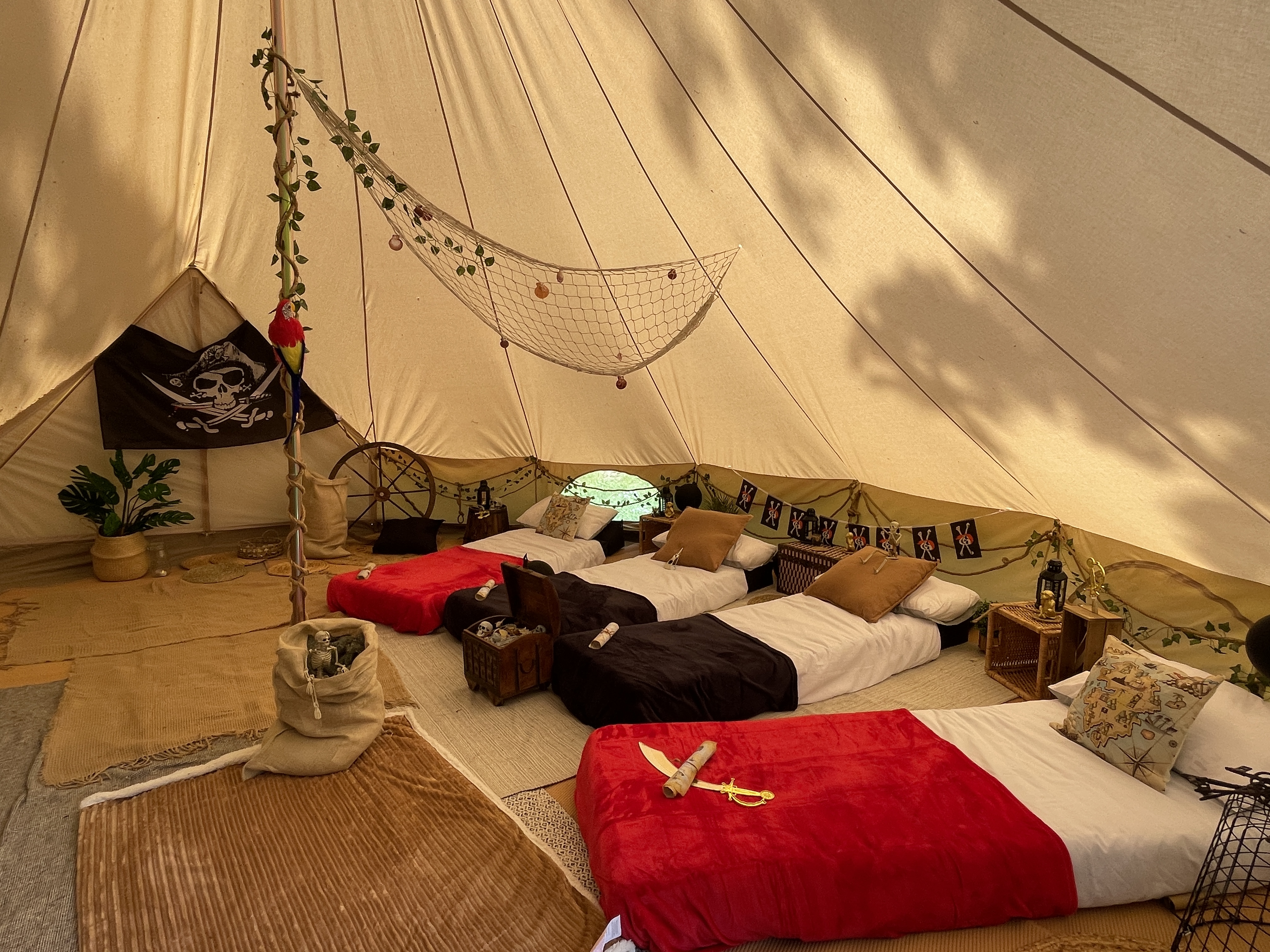 Inside a spacious tent, three neatly arranged beds feature crisp white linens and vibrant red and black throws, with a playful pirate theme, accented by lush greenery and warm golden lighting, creating a cozy, adventurous atmosphere.