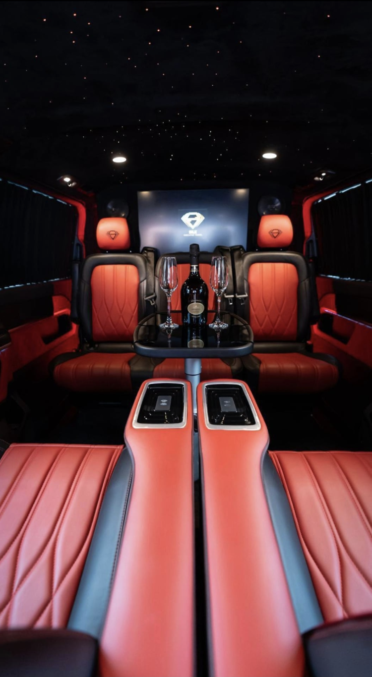 Luxurious limo interior with red quilted leather seats, black glass table, champagne flutes and bottle, starry ceiling, moody ambient lighting.