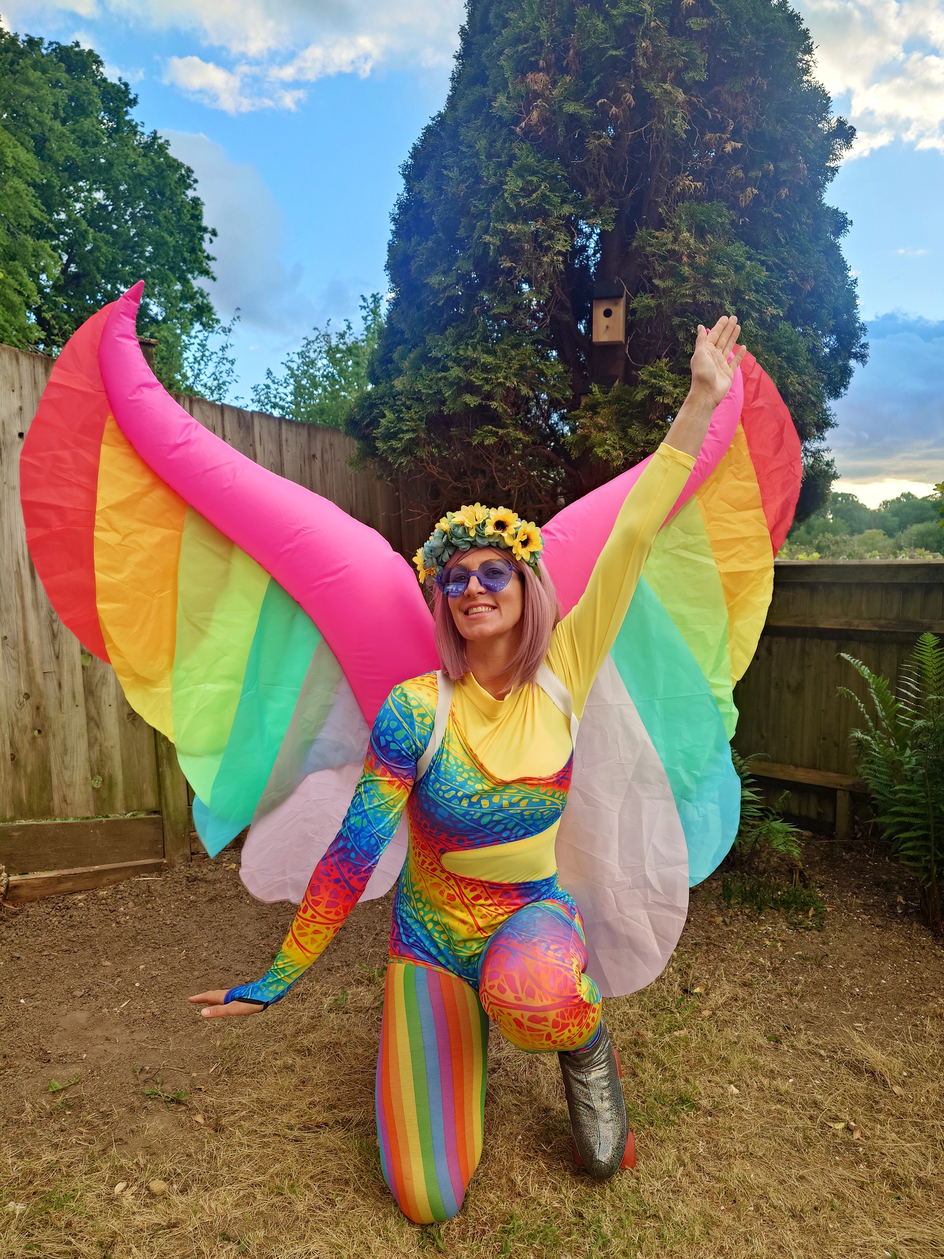 A performer in a shimmering silver outfit holds a glowing hoop above her head, posed dramatically on a sunny grassy field, showcasing vibrant energy.