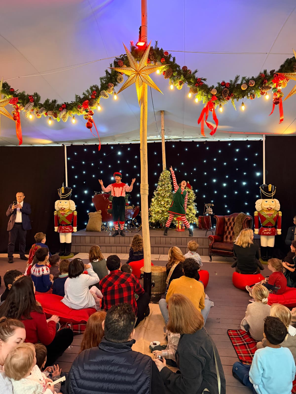 A vibrant holiday scene features performers and children captivated by a festive show, adorned with twinkling lights and charming decorations.