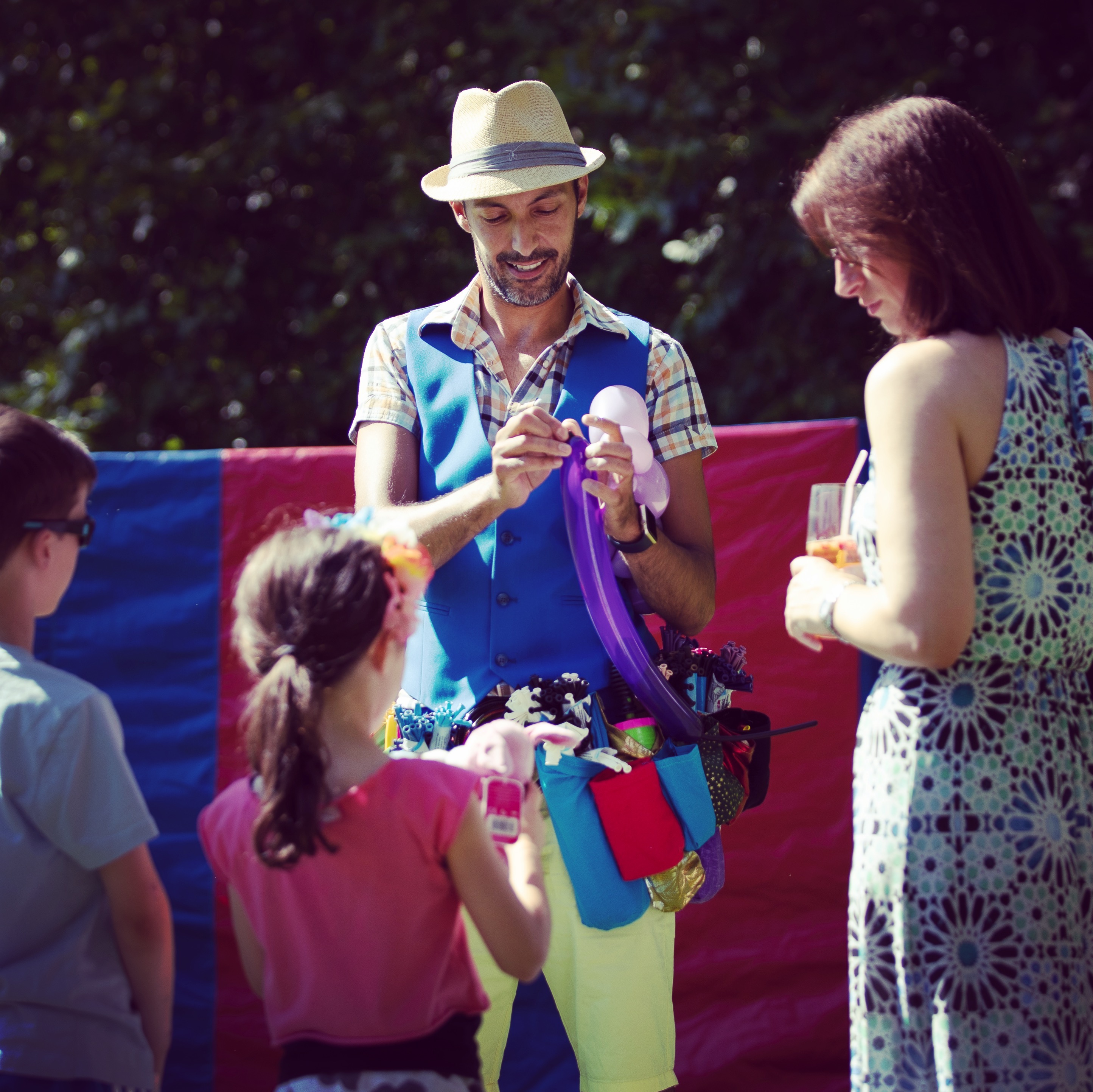 A cheerful entertainer in a bright blue vest creates balloon art for children, surrounded by greenery and colorful fabric backdrops under warm sunlight.