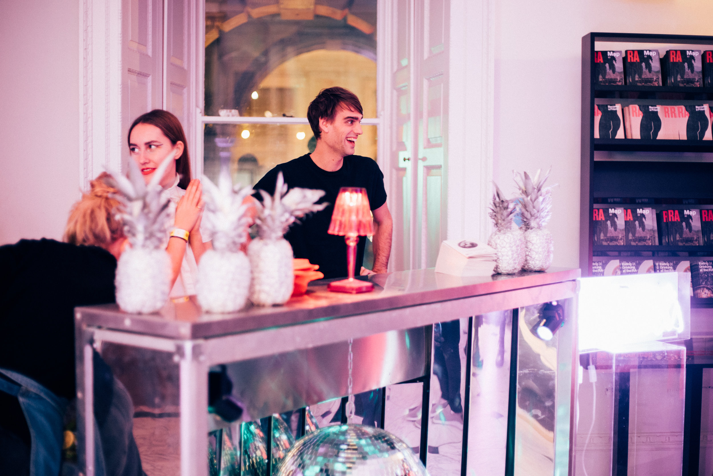 A modern catering setup features a shiny metal bar with decorative pineapples and a vibrant pink lamp, illuminated by soft purple lighting.