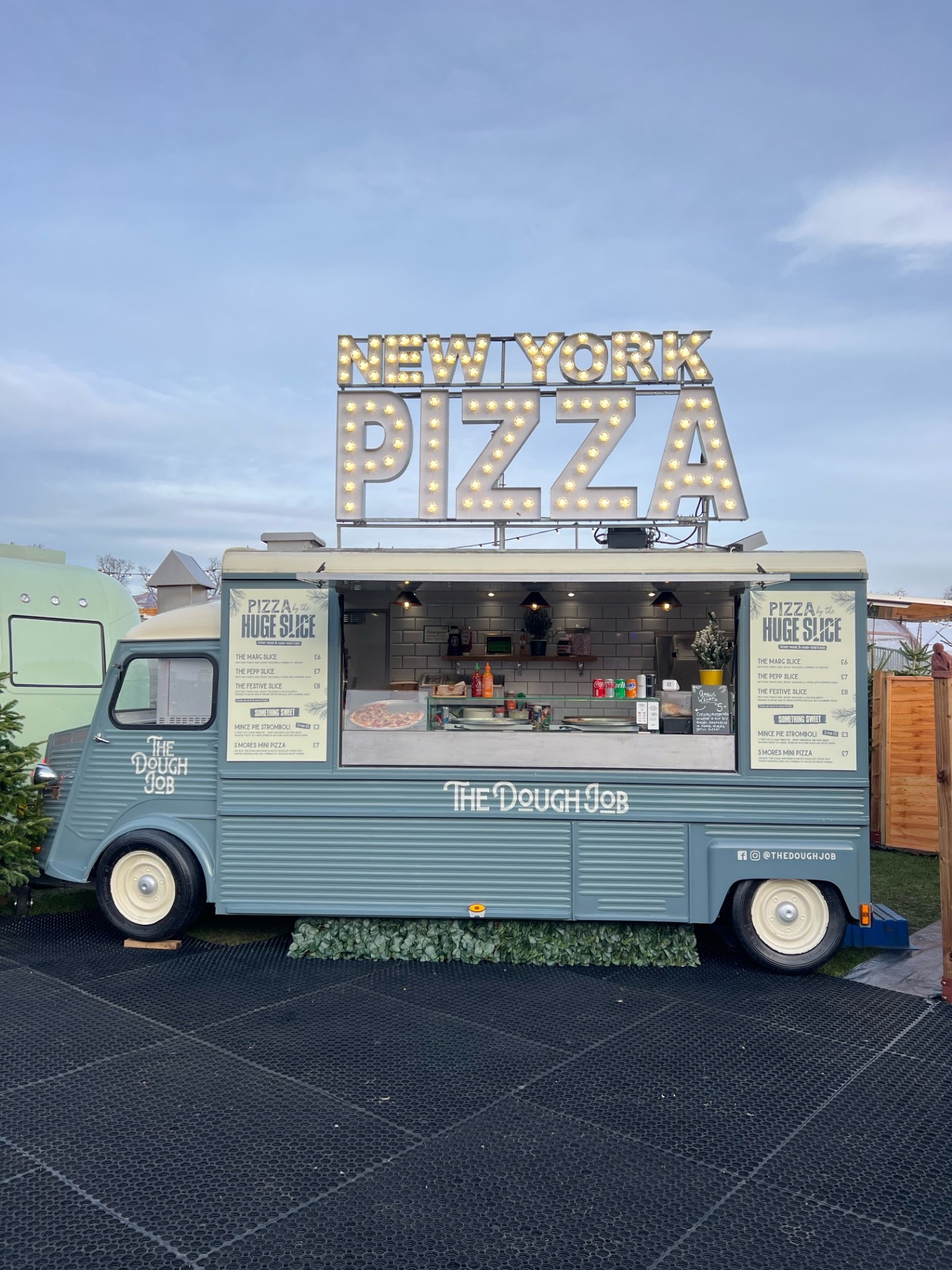 Blue vintage pizza catering van “The Dough Job” with glowing NEW YORK PIZZA sign, polished open serving window, and tidy evening setup.