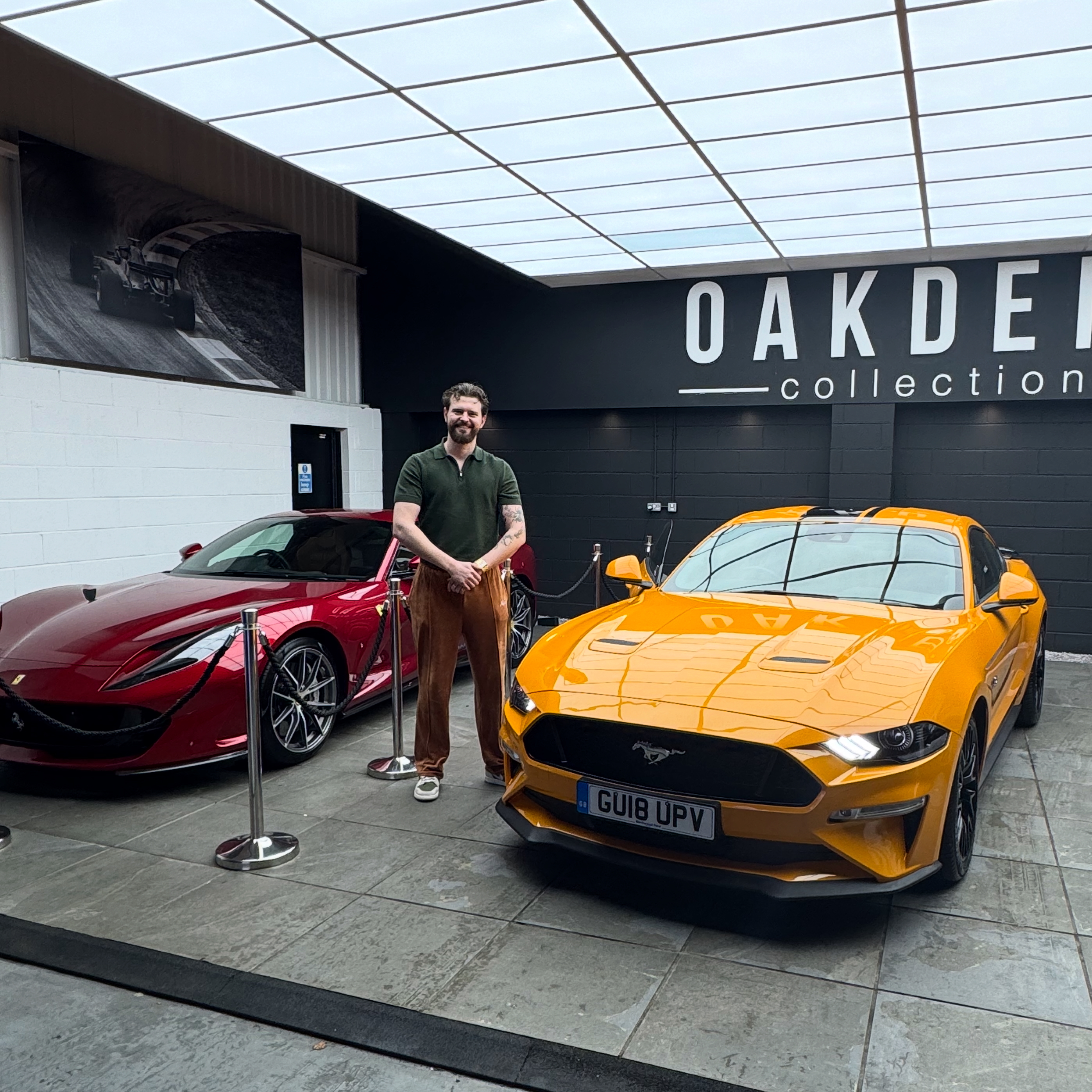 In a sleek showroom, a stylish man stands between a red Ferrari and an orange Mustang, showcasing vibrant vehicles under bright lights.