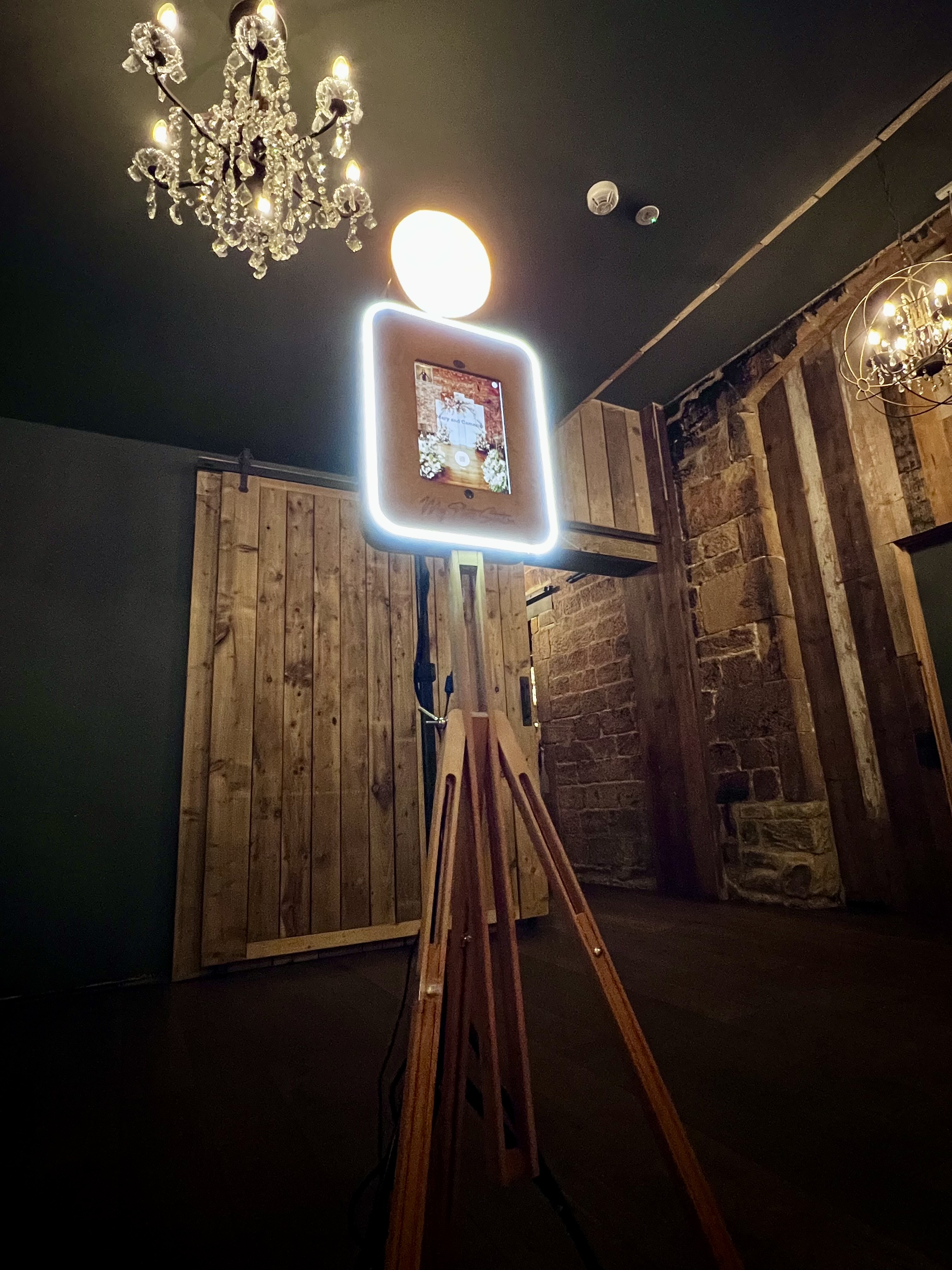 A stylish wooden tripod holds a glowing photo booth, illuminated by a chic chandelier against rustic wooden walls, creating a vibrant ambiance.