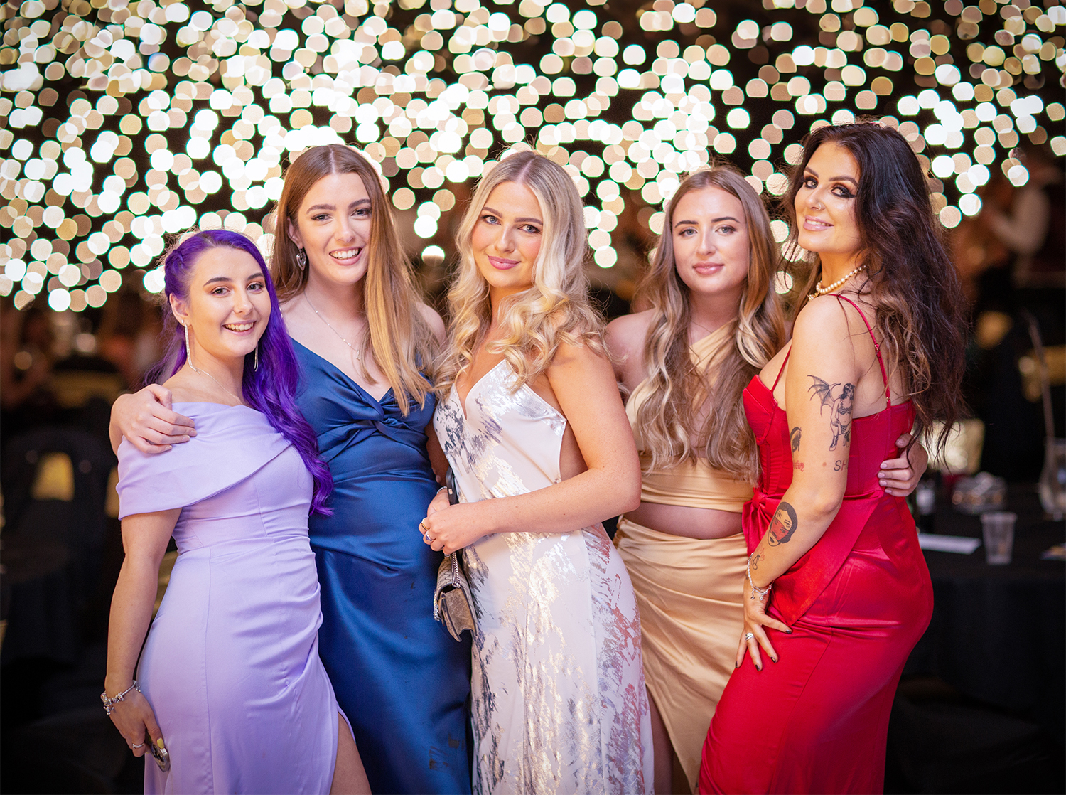 Five elegantly dressed women pose together, glowing under warm fairy lights, embodying celebration and camaraderie at a festive event.