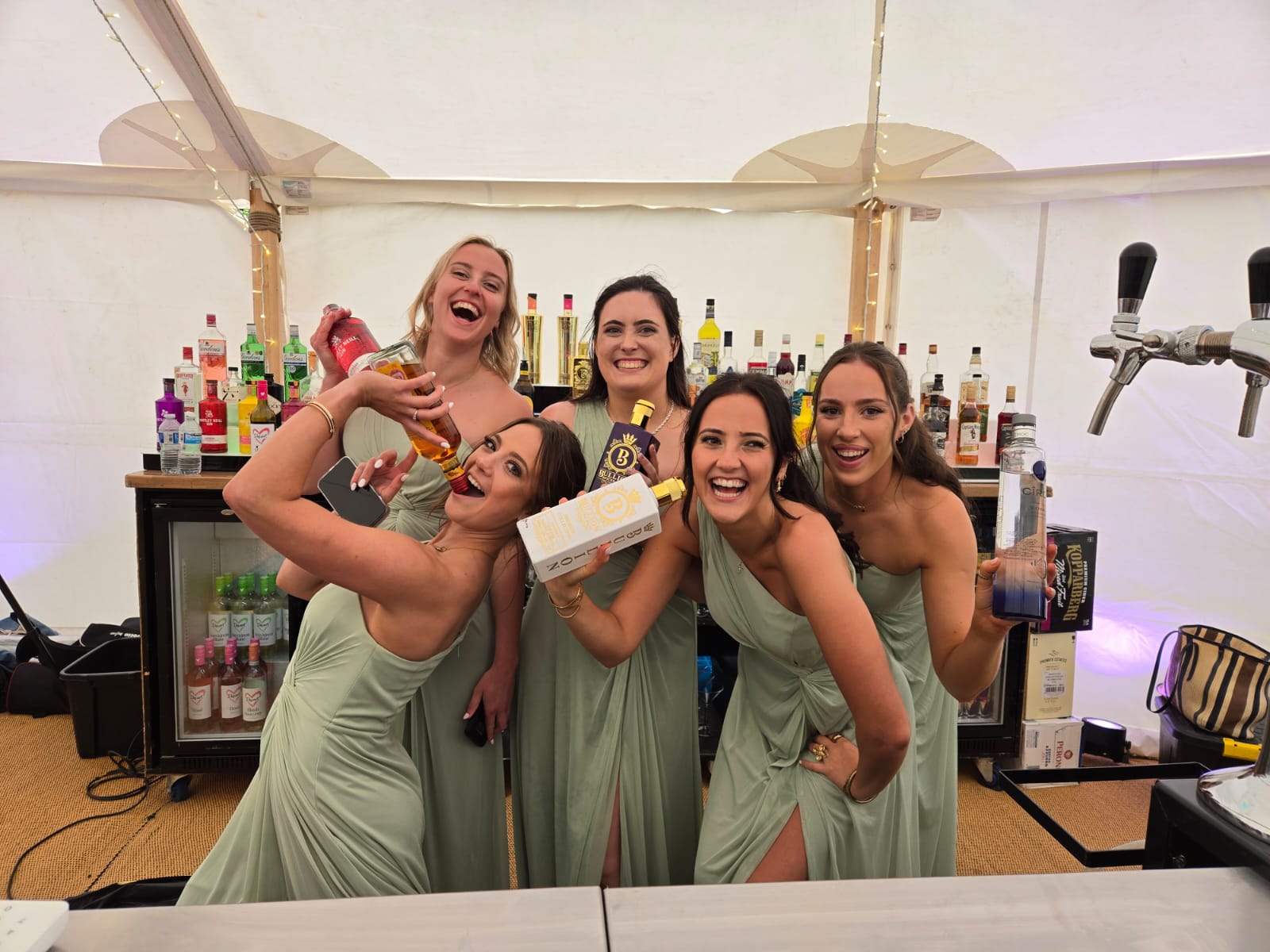 Five smiling bartenders in sage gowns pose at a polished bar, with liquor bottles, taps, and warm fairy lights in a chic catering tent.