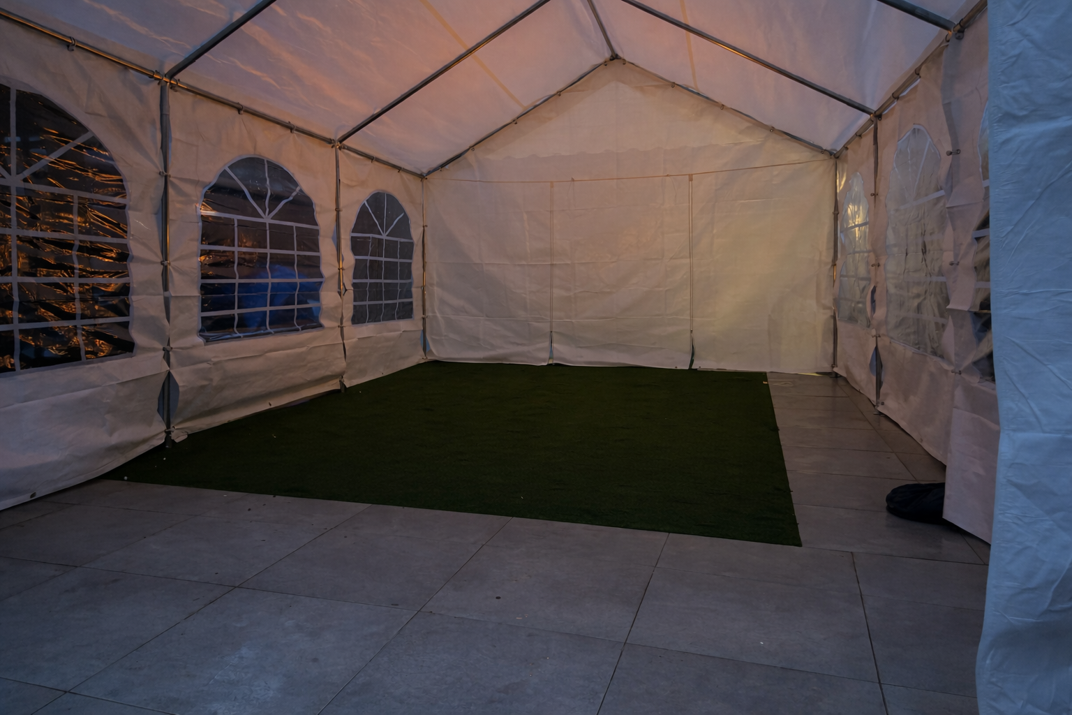 A spacious white tent features large arched windows, with artificial grass and stone flooring, creating a clean, airy atmosphere ideal for catering events.