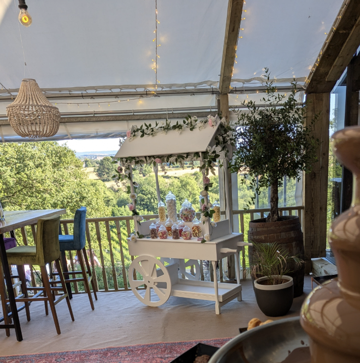 A charming candy cart with colorful jars sits under soft string lights, surrounded by rustic wooden decor and lush greenery, creating a warm atmosphere.