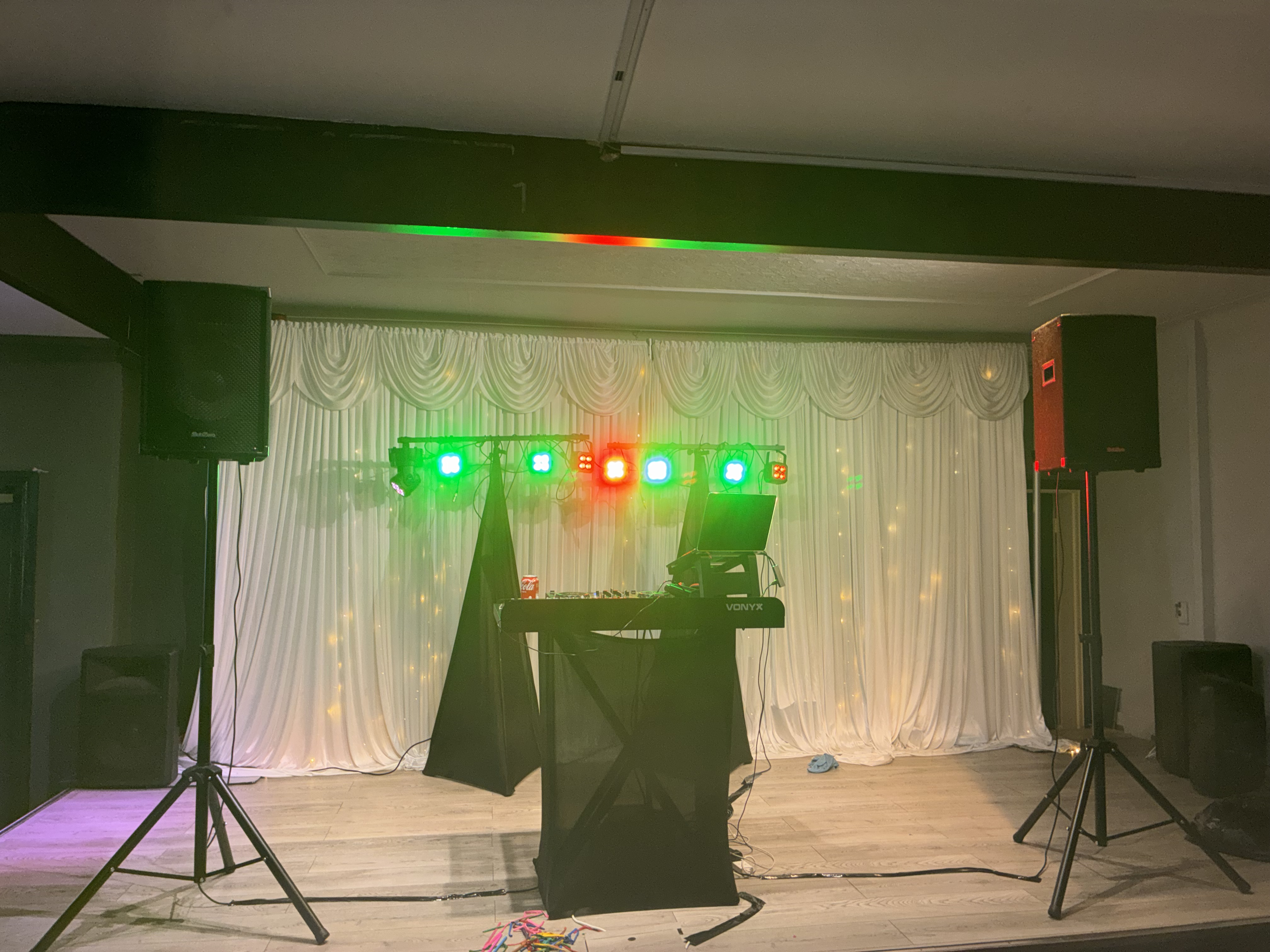 Empty DJ stage with green and red LED lights, twin speakers, laptop, and Vonyx console before white draped curtains on wood flooring.