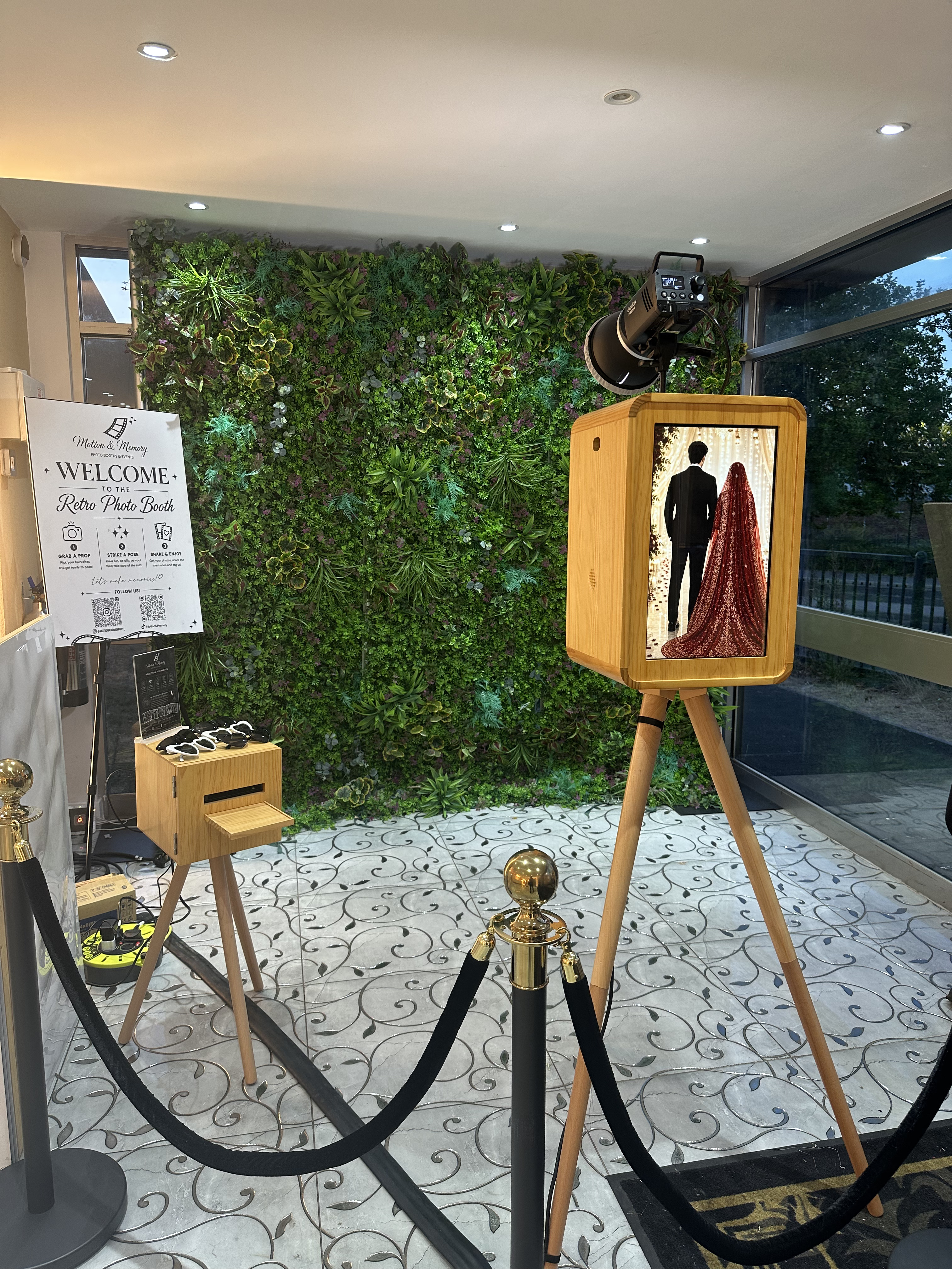 Elegant photo booth setup with retro wood kiosks, green wall backdrop, polished gold stanchions, bright indoor lighting, and crisp event signage.