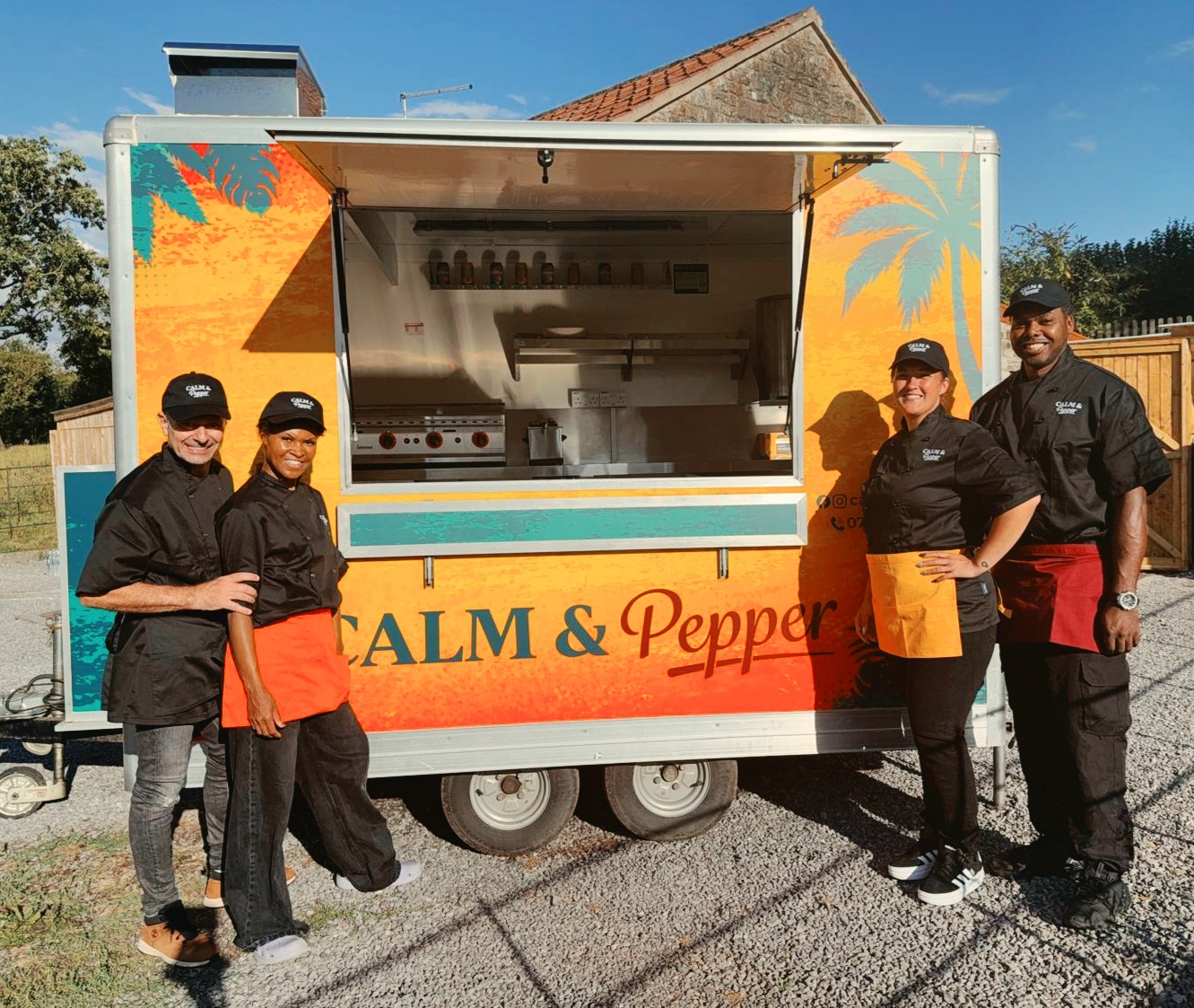 Bright daylight frames a colorful Calm & Pepper catering food truck, with four smiling chefs in black uniforms posed beside the open, polished stainless-steel service window.