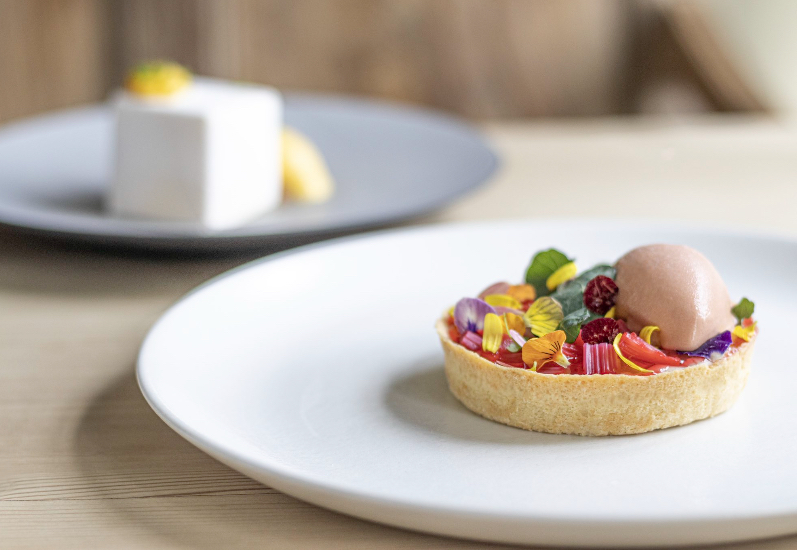 A vibrant tart with edible flowers and a berry scoop sits on a sleek white plate, exuding elegance under soft, natural lighting.