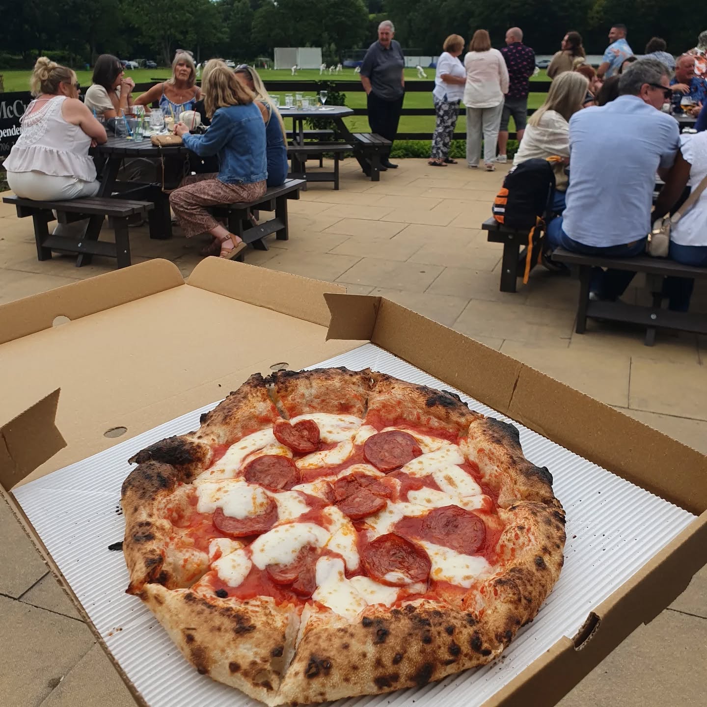 A fresh pepperoni pizza with melted mozzarella is showcased in a box, amidst a lively outdoor setting with picnic-style seating and guests enjoying a sunny afternoon.