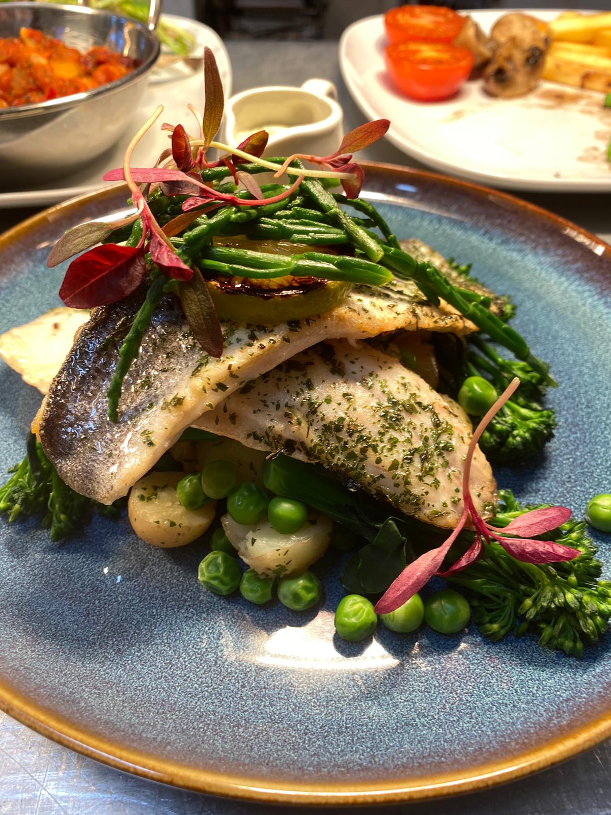 Elegant catering plate with herb-crusted fish, asparagus, peas and potatoes, artfully stacked on blue ceramic under warm buffet lighting.