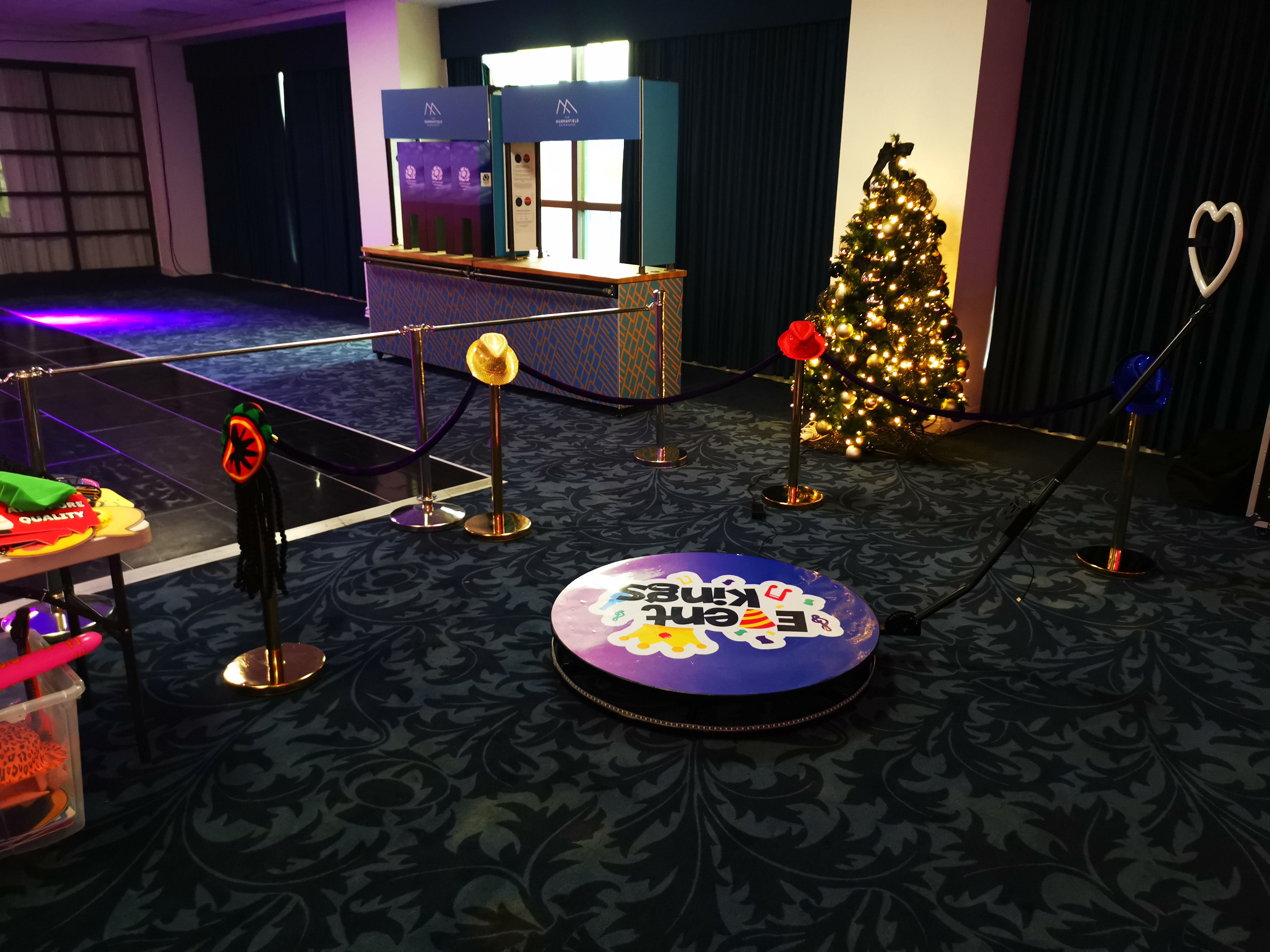The vibrant space features a decorated Christmas tree beside a colorful photo backdrop, with playful props arranged on a black table. Soft purple lighting creates a festive atmosphere, perfect for a lively event.