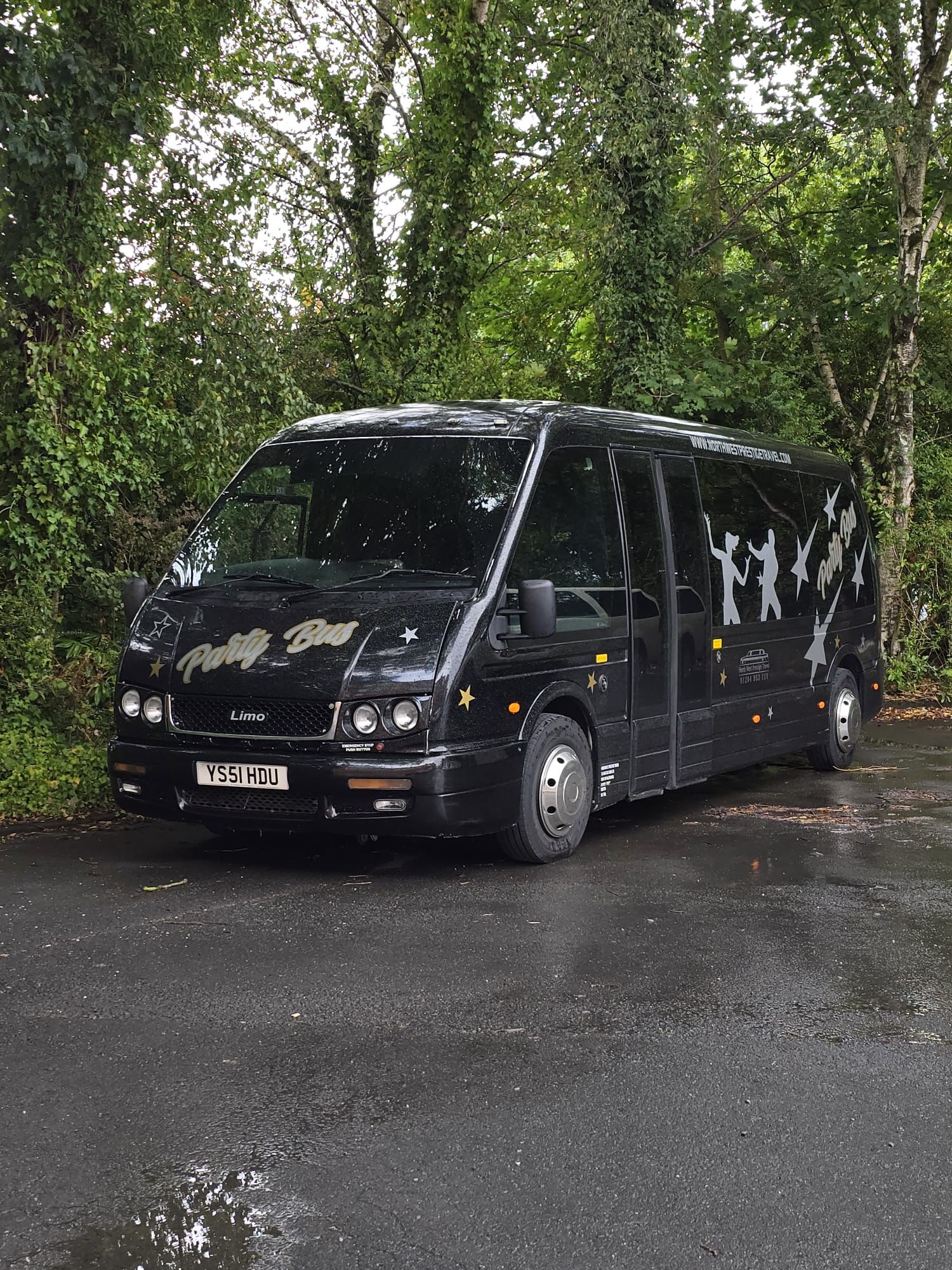 Black party bus limo with gold stars and dancing silhouettes, parked on wet asphalt amid lush trees, sleek catering-style transport.