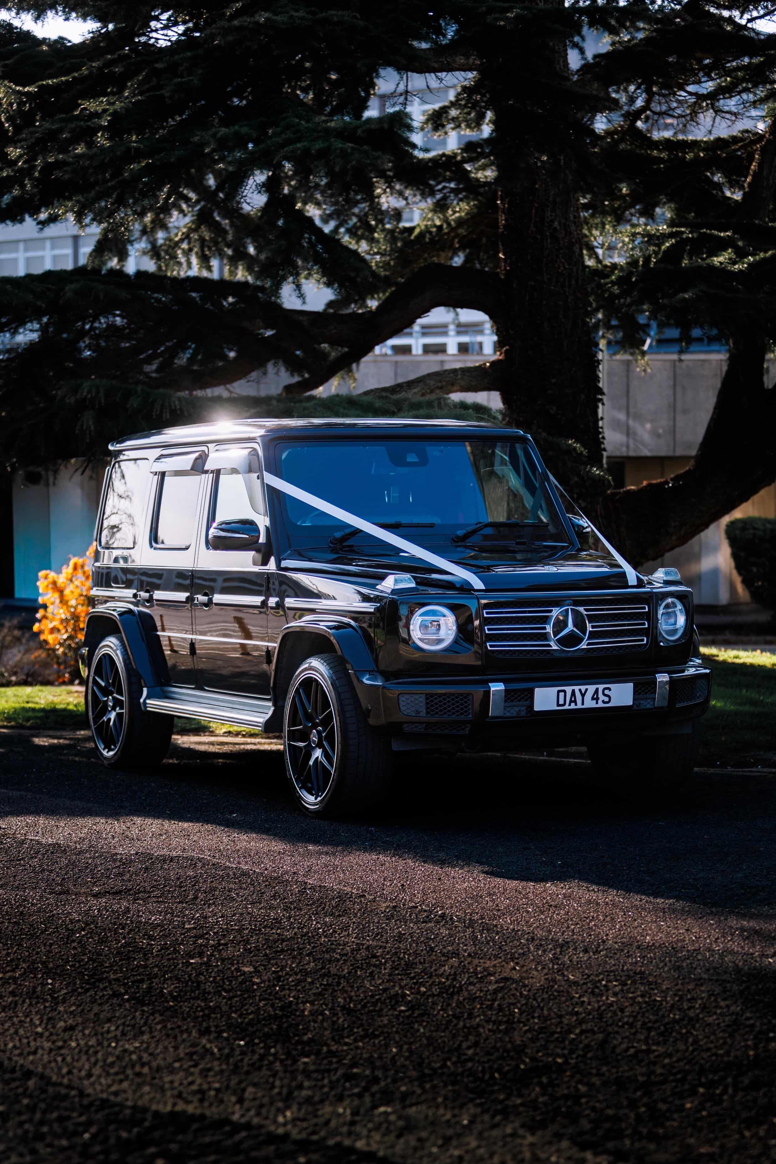 A sleek black Mercedes G-Class sits parked, wrapped with a white ribbon under bright sunlight, showcasing its luxury design against lush greenery.