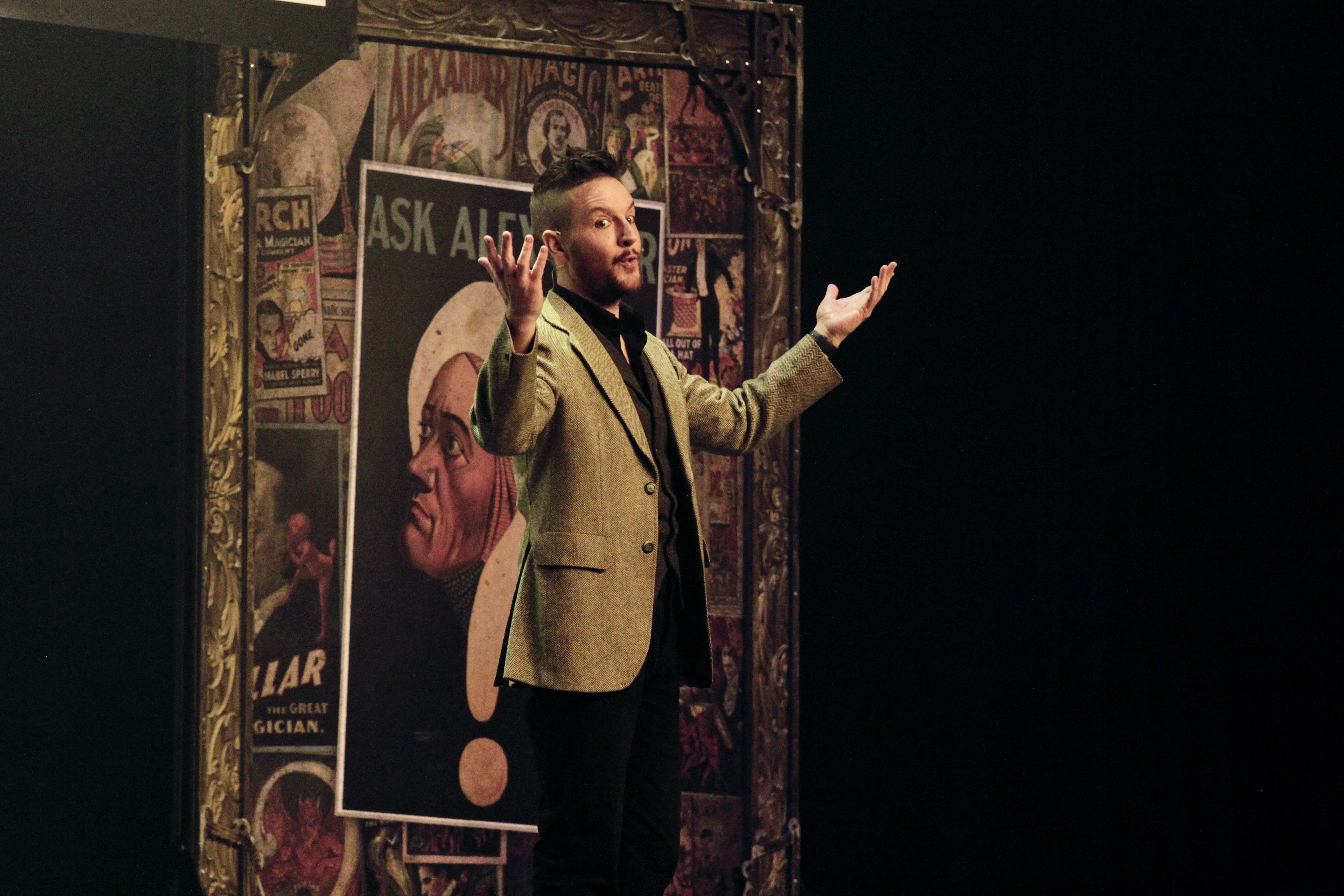 A performer in a vintage-style blazer gestures animatedly against a backdrop of retro magician posters, creating an engaging atmosphere.