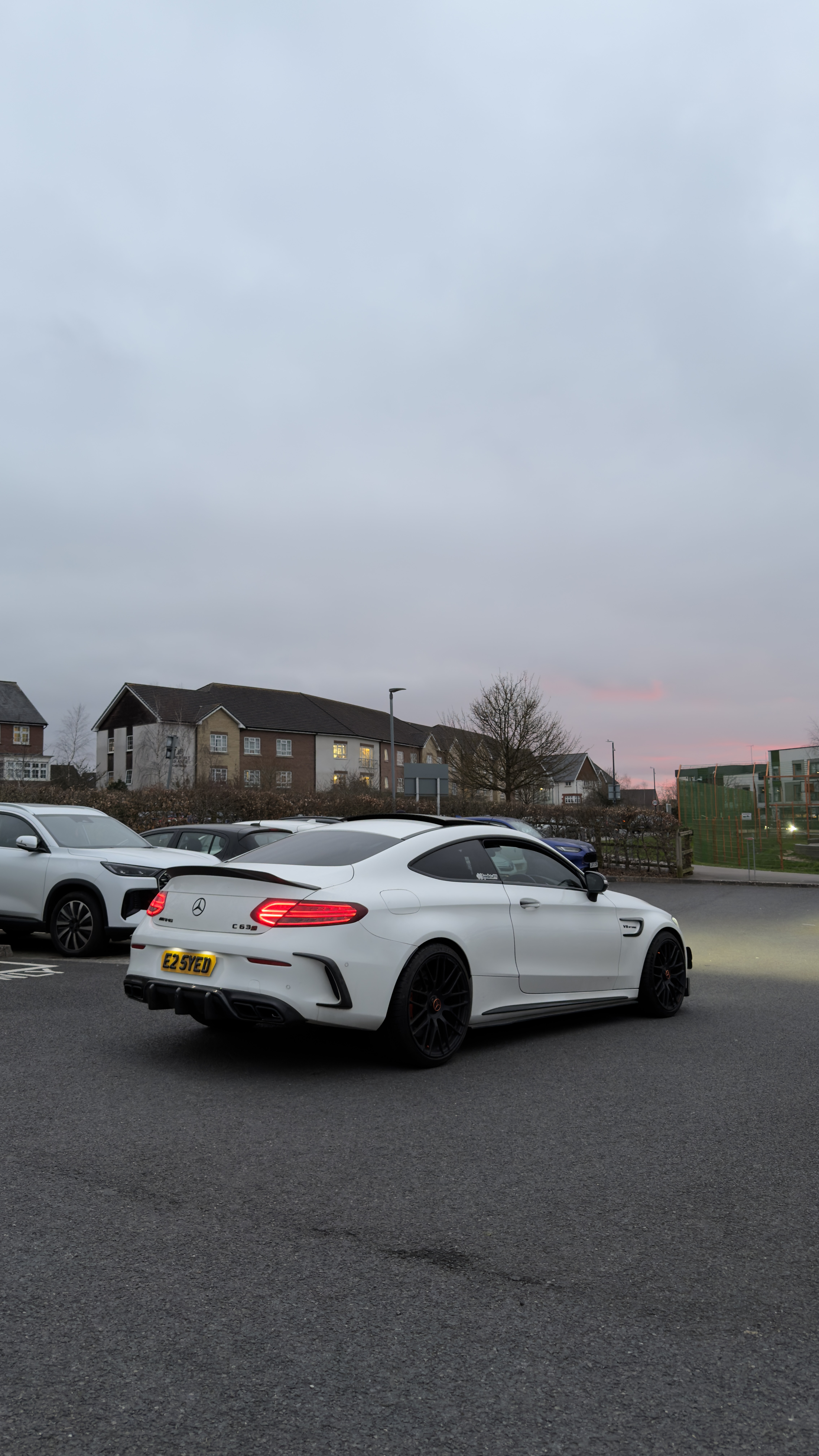 A sleek white Mercedes AMG C63 stands out against gray skies in a parking lot, showcasing its sporty design and vibrant taillights.