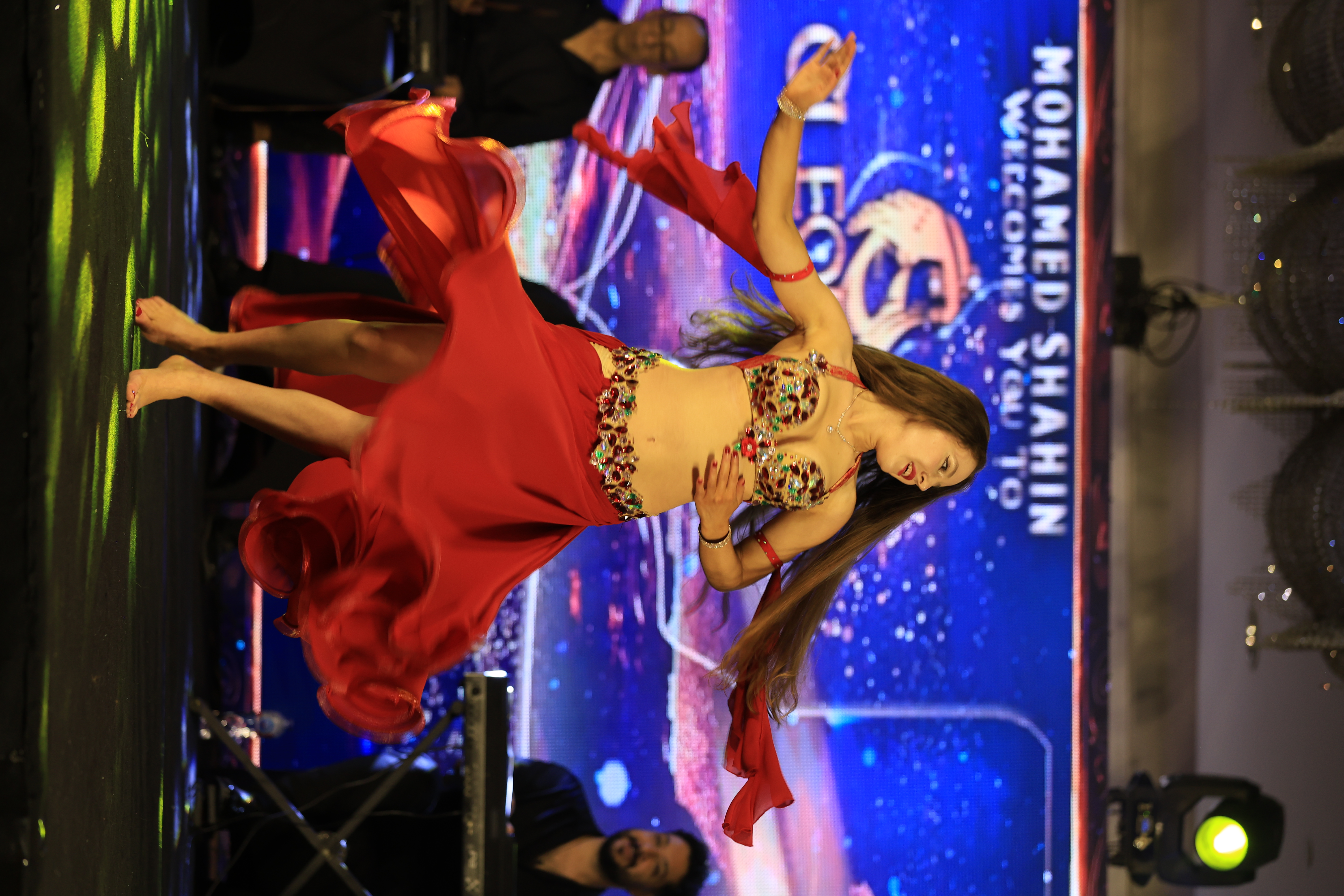 A belly dancer in a jeweled red costume spins onstage under blue LED backdrop, with live musicians and event lighting.