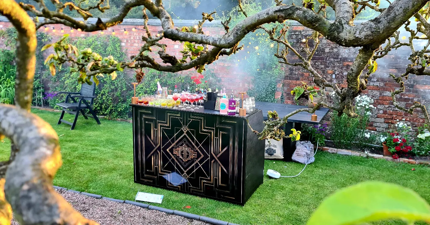 A sleek black bar with art deco design displays colorful cocktails in a lush garden, framed by blooming flowers and soft evening light.