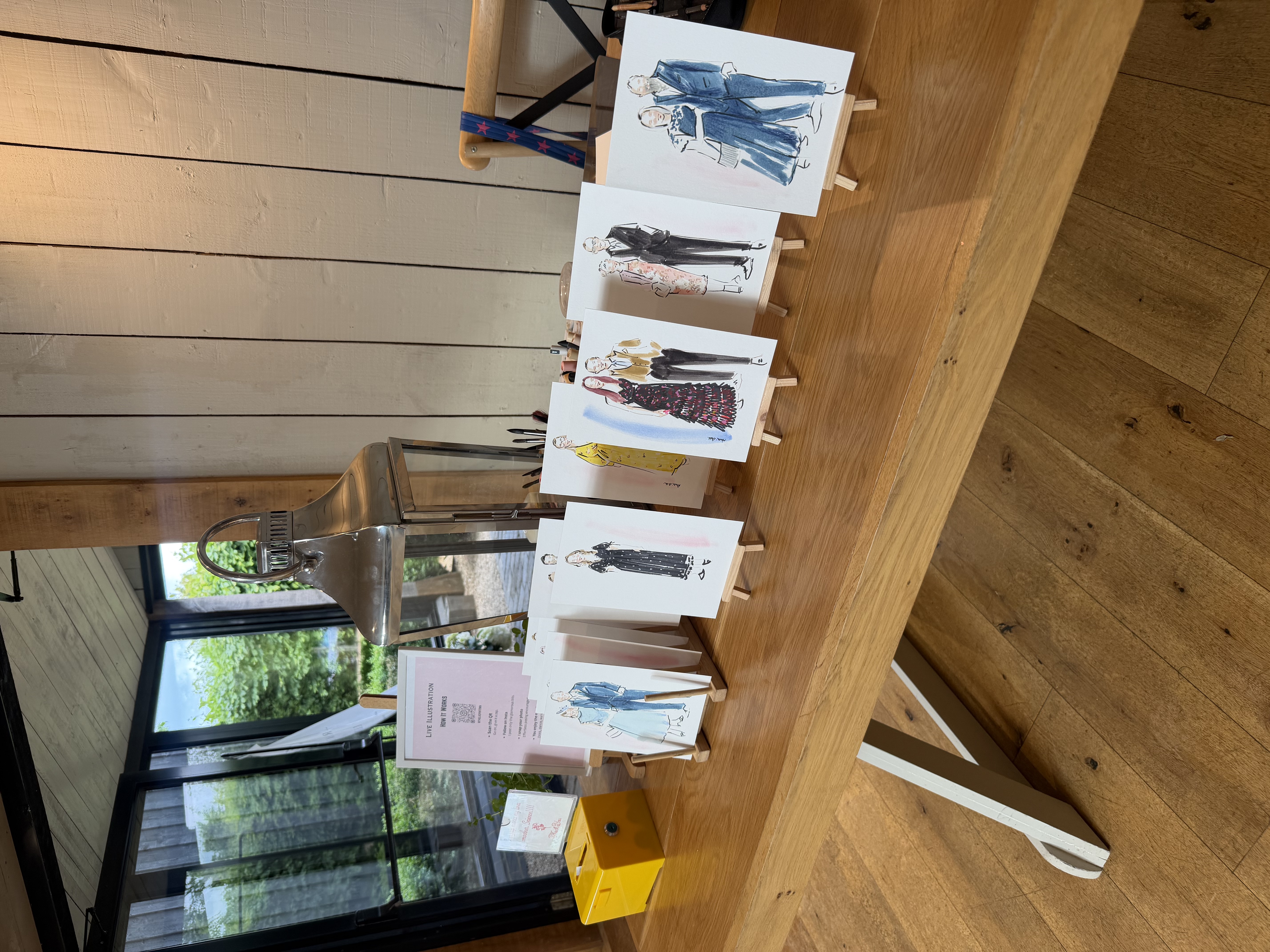 Elegant live illustration display on a rustic wood table, with easels, polished lantern, and wedding sketches in soft daylight.