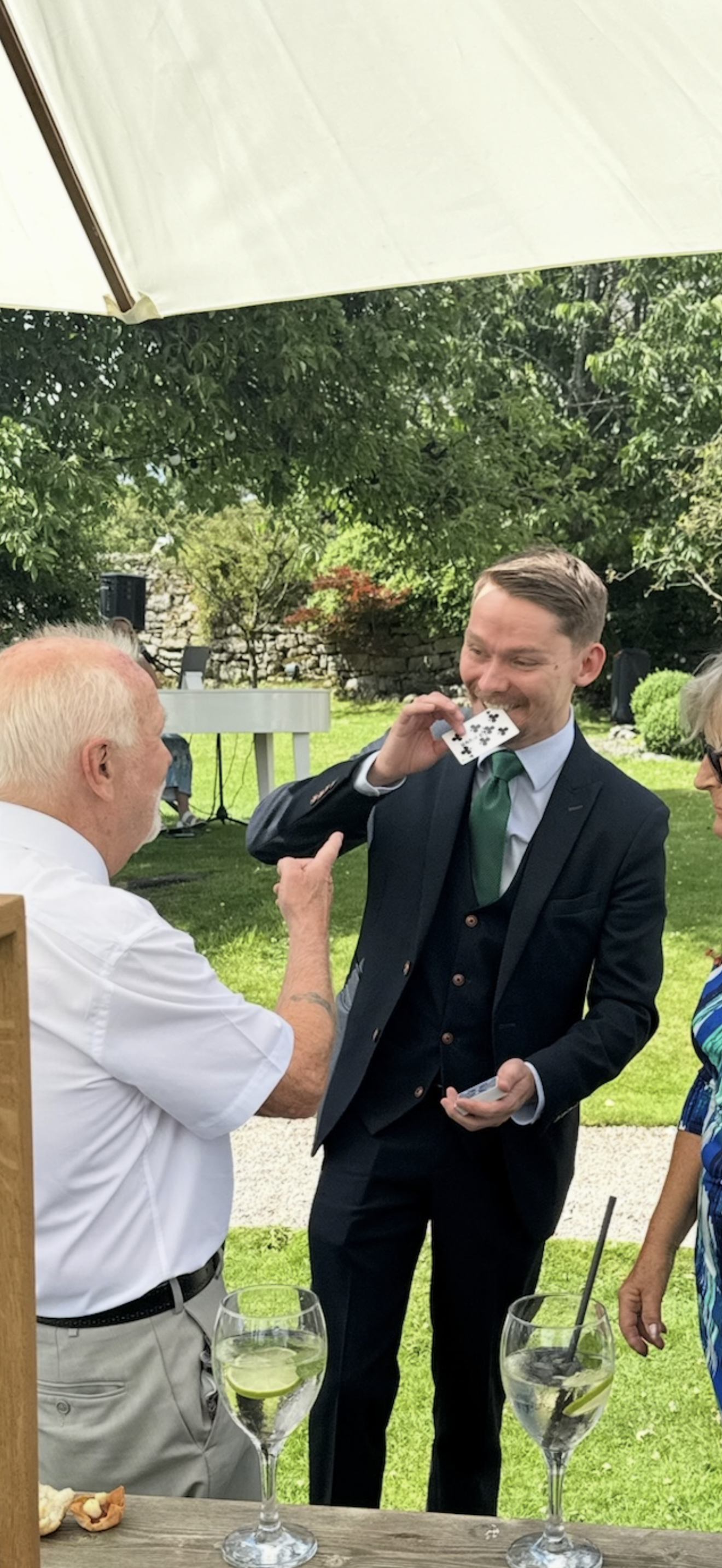 Outdoor cocktail catering with a magician in a black suit, lemon spritz glasses on rustic wood, under bright garden daylight.