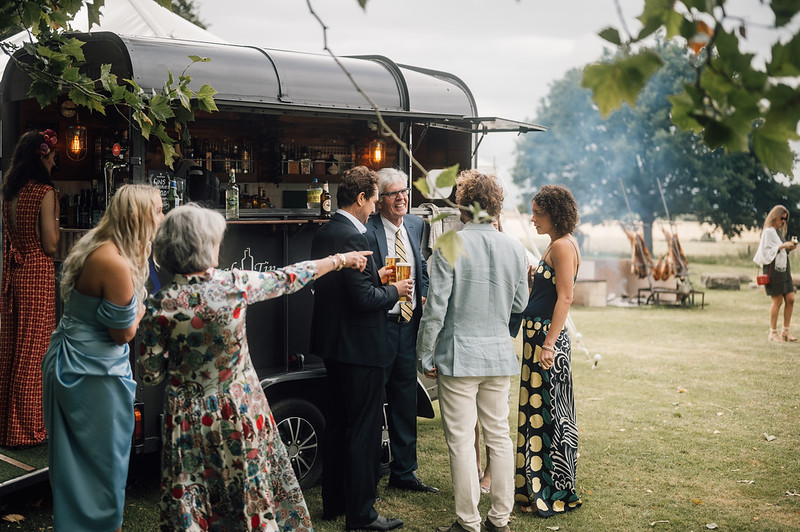 A vintage bar trailer serves cocktails at an outdoor gathering, where elegantly dressed guests mingle in soft afternoon light, creating a festive vibe.
