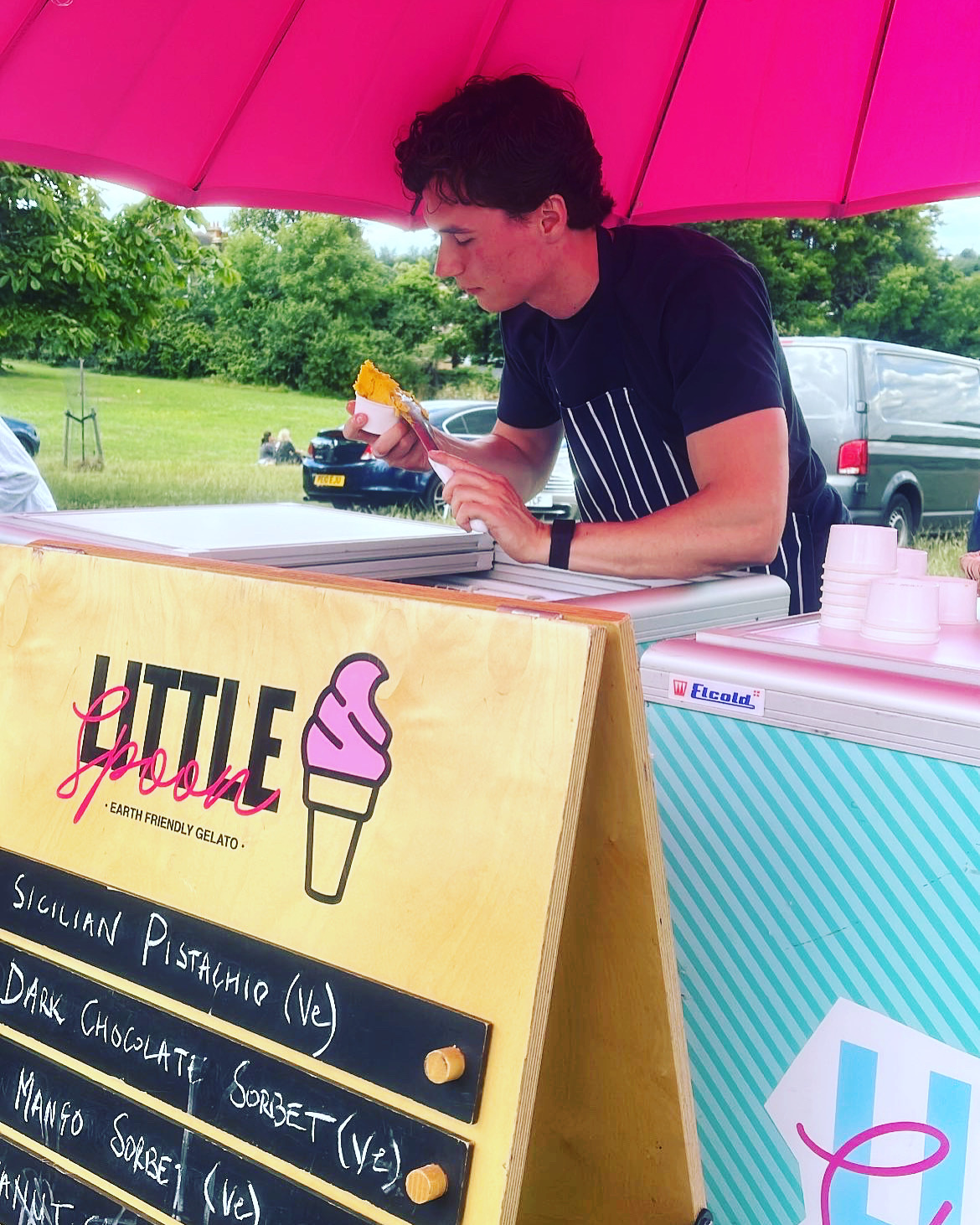 A young man serves vibrant gelato under a bright pink umbrella; the wooden sign lists flavors like Sicilian Pistachio and Mango Sorbet.