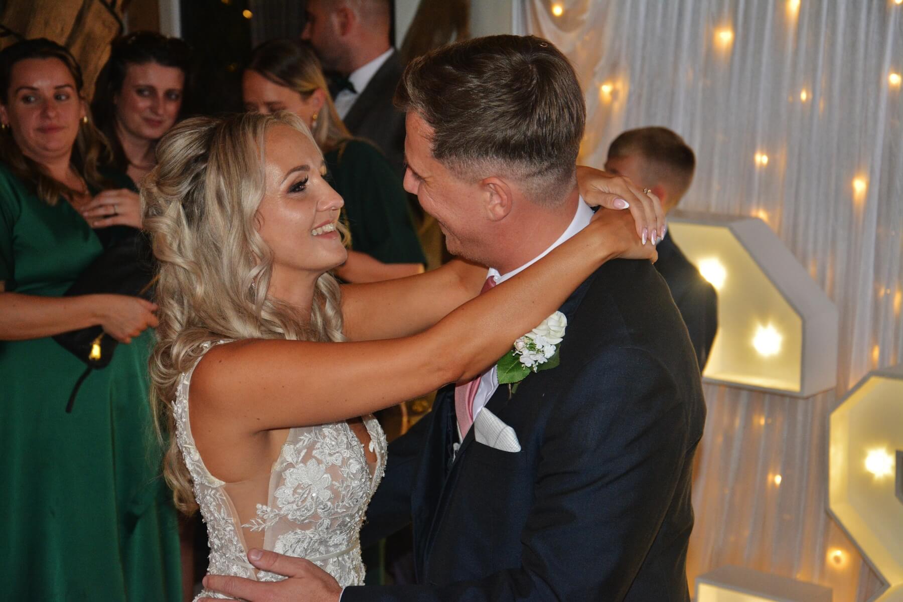 Bride and groom dance in warm fairy lights, surrounded by bridesmaids in green at an elegant wedding reception, polished and intimate.