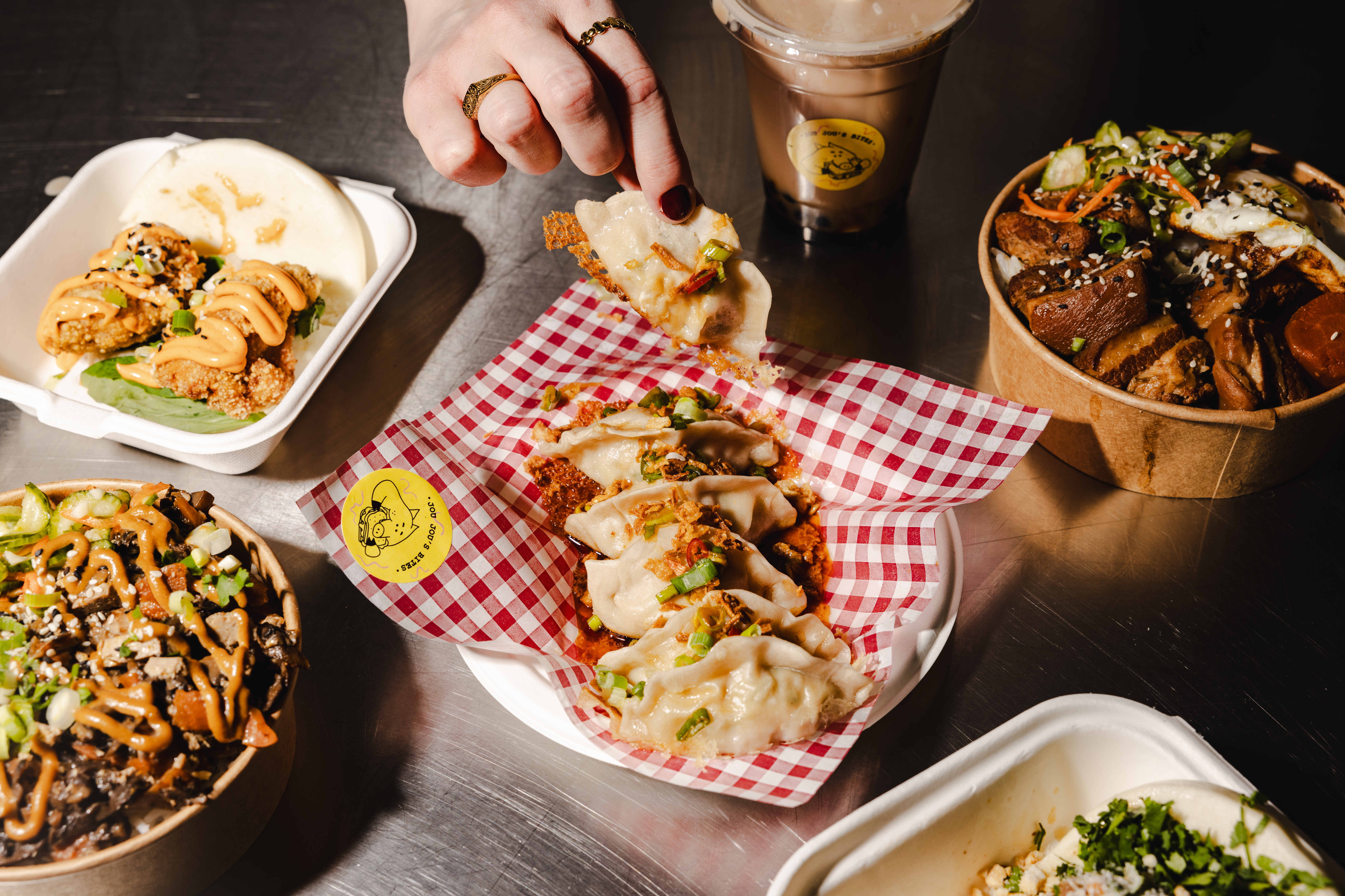 A hand reaches for a dumpling atop a vibrant red-checkered paper amid spread-out dishes, creating a lively, inviting atmosphere.