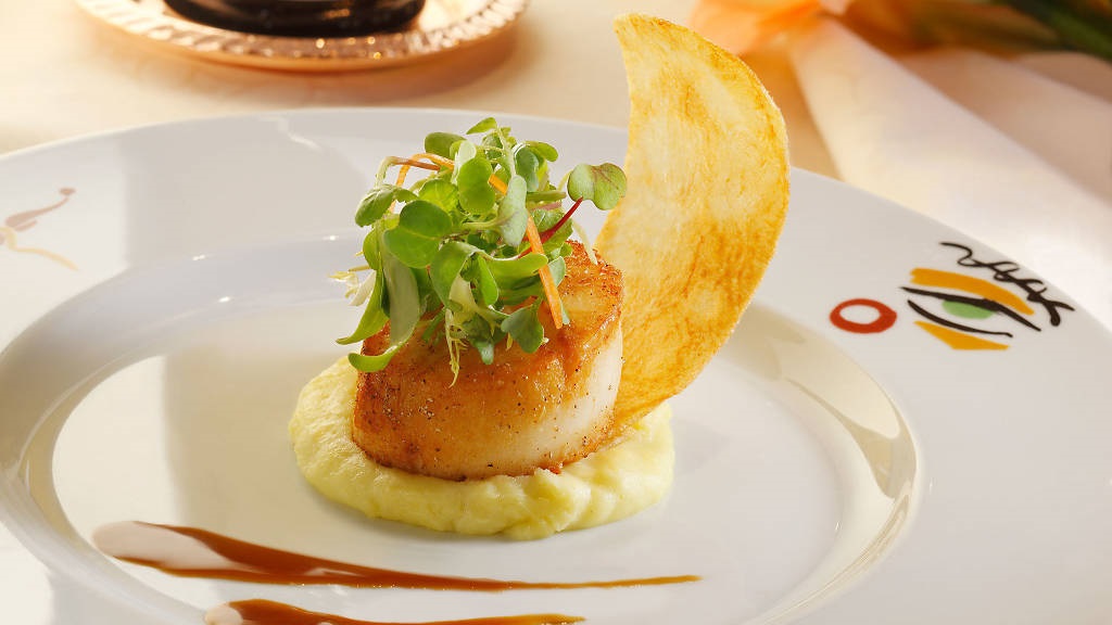 A beautifully plated scallop sits atop creamy potato puree, garnished with microgreens, with a crisp chip leaning elegantly, all in warm lighting.