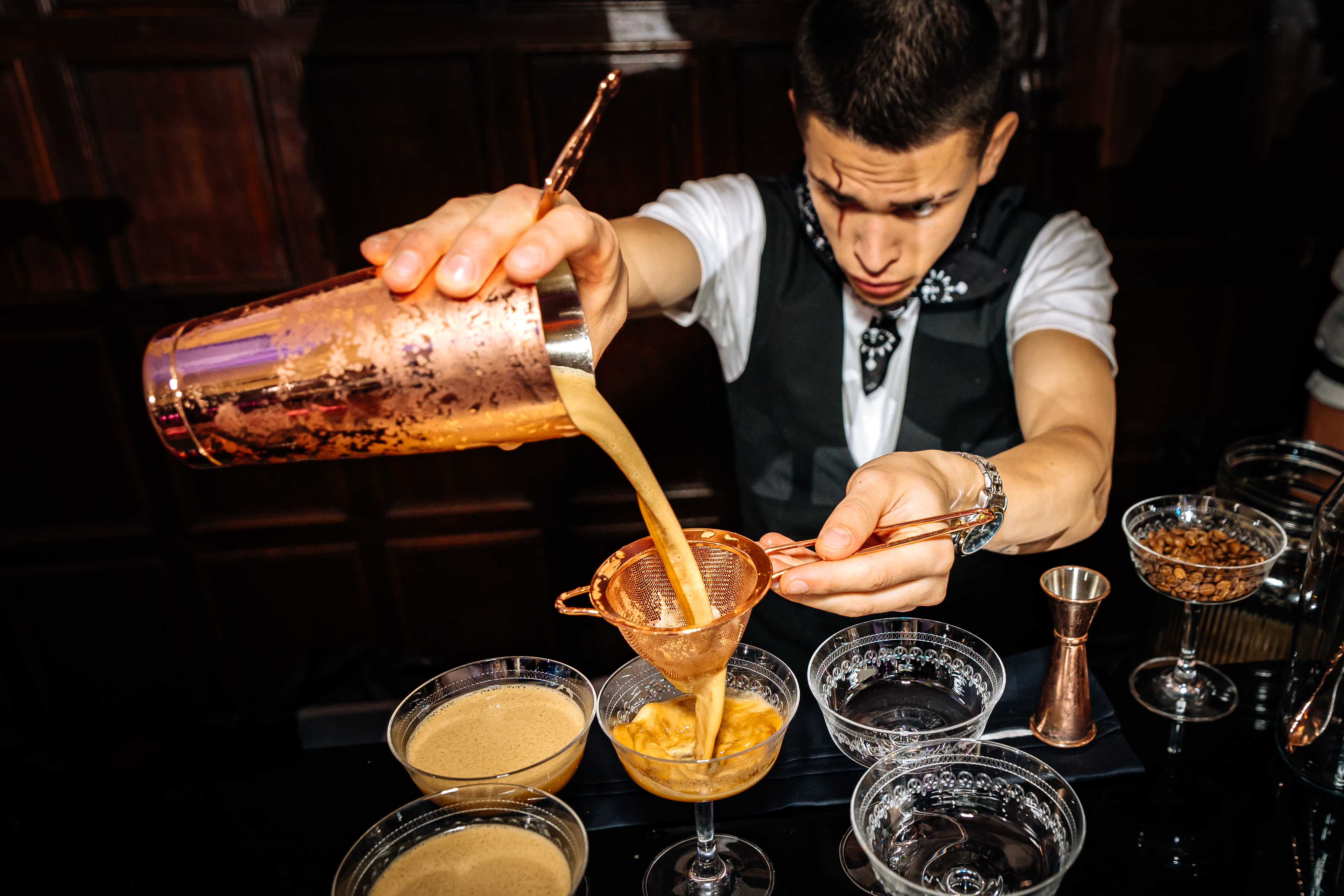 A bartender in a sleek black vest carefully pours a creamy beverage through a copper strainer into elegant glassware, lit by warm ambiance.
