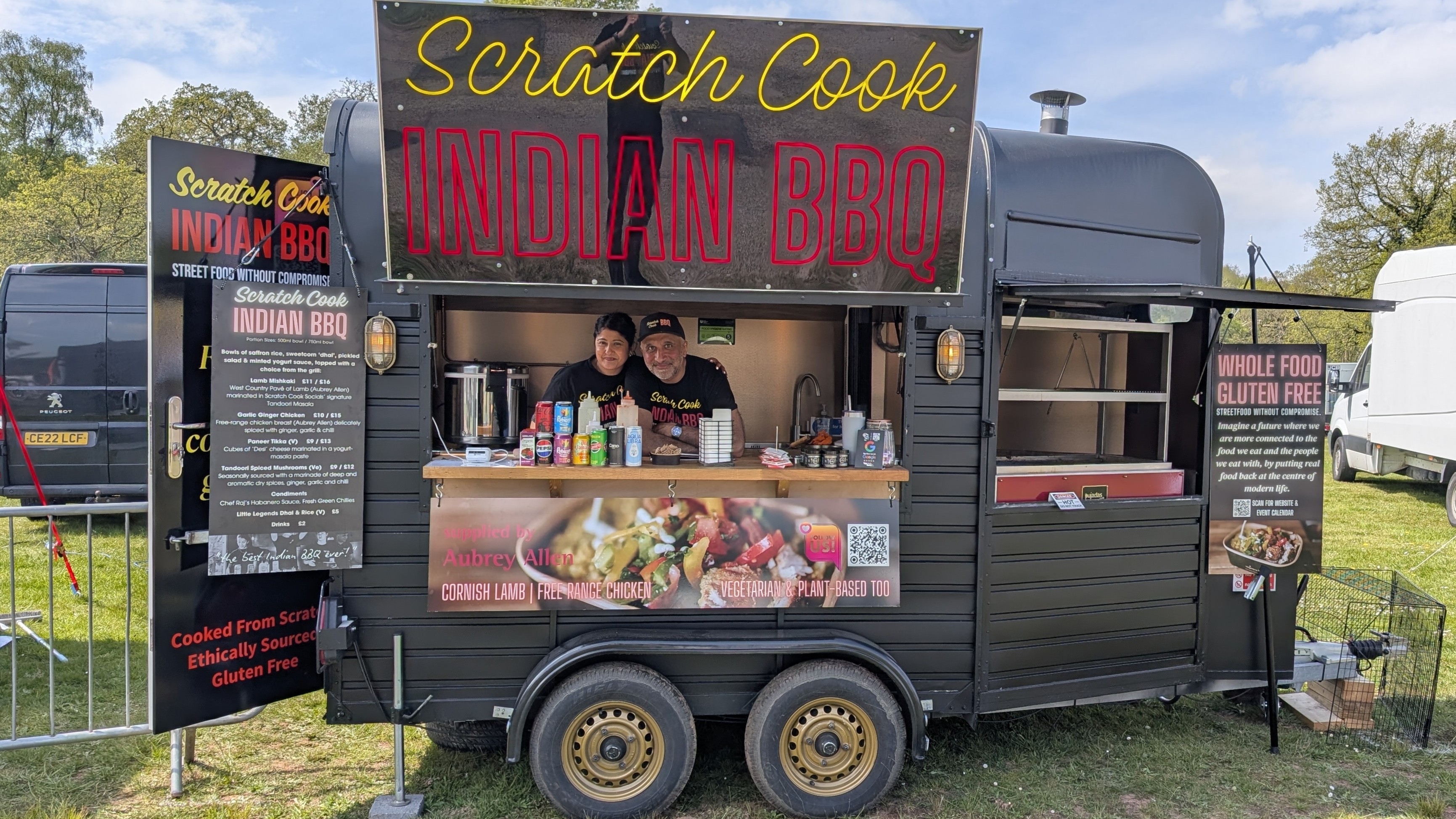 Bright daylight on a black catering trailer serving Scratch Cook Indian BBQ, with colorful menu boards, polished counter, and a lively street-food vibe.