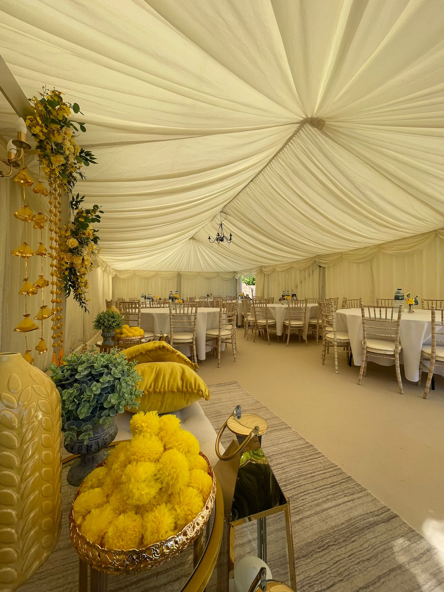 A bright, elegant tent features crisp white drapery and gold accents; styled tables with silver chiavari chairs are adorned with vibrant yellow marigolds.