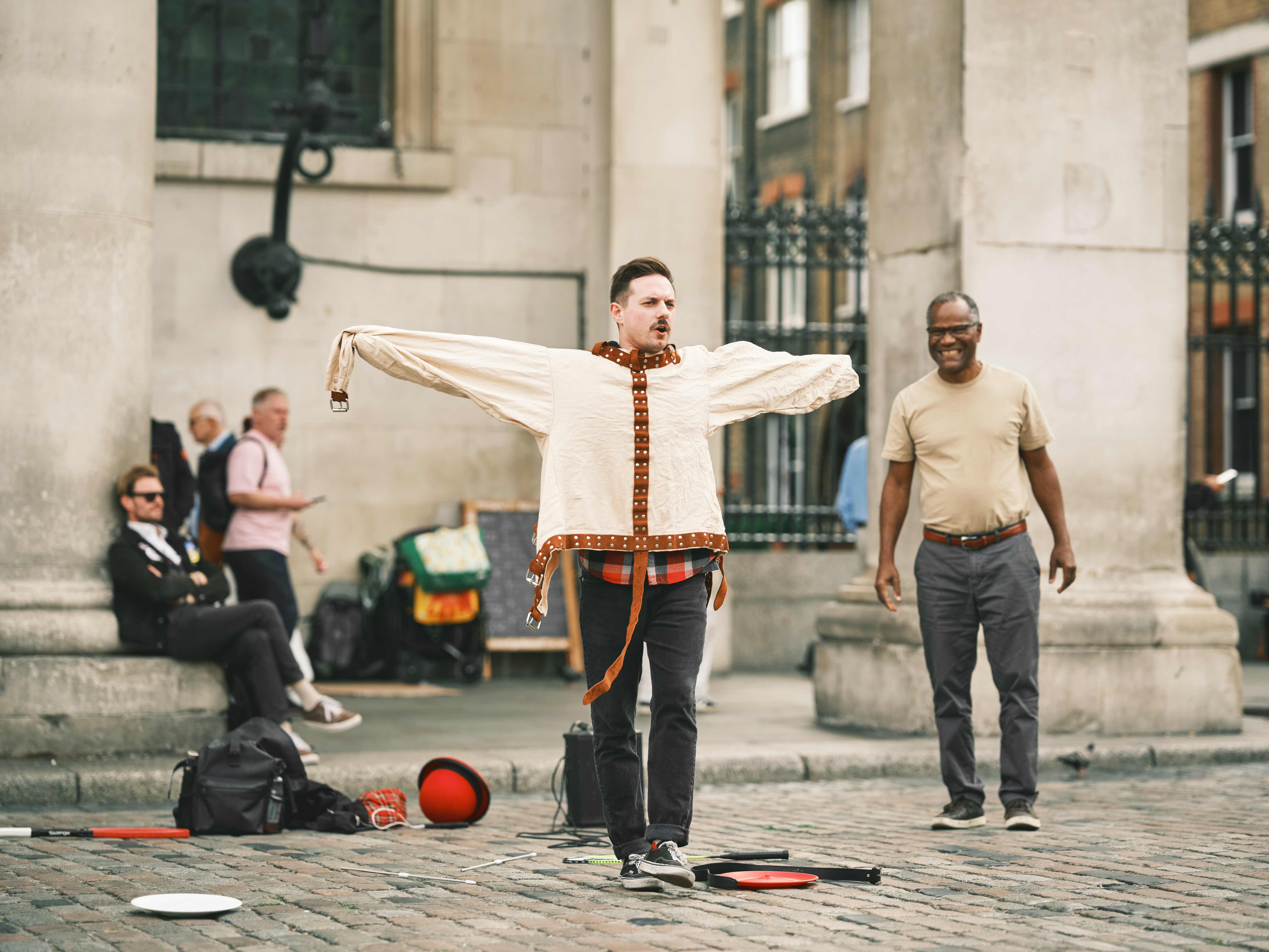 Street performer in a cream tunic and leather harness stands on cobblestones, arms outstretched, with props and onlookers by stone columns in soft daylight.