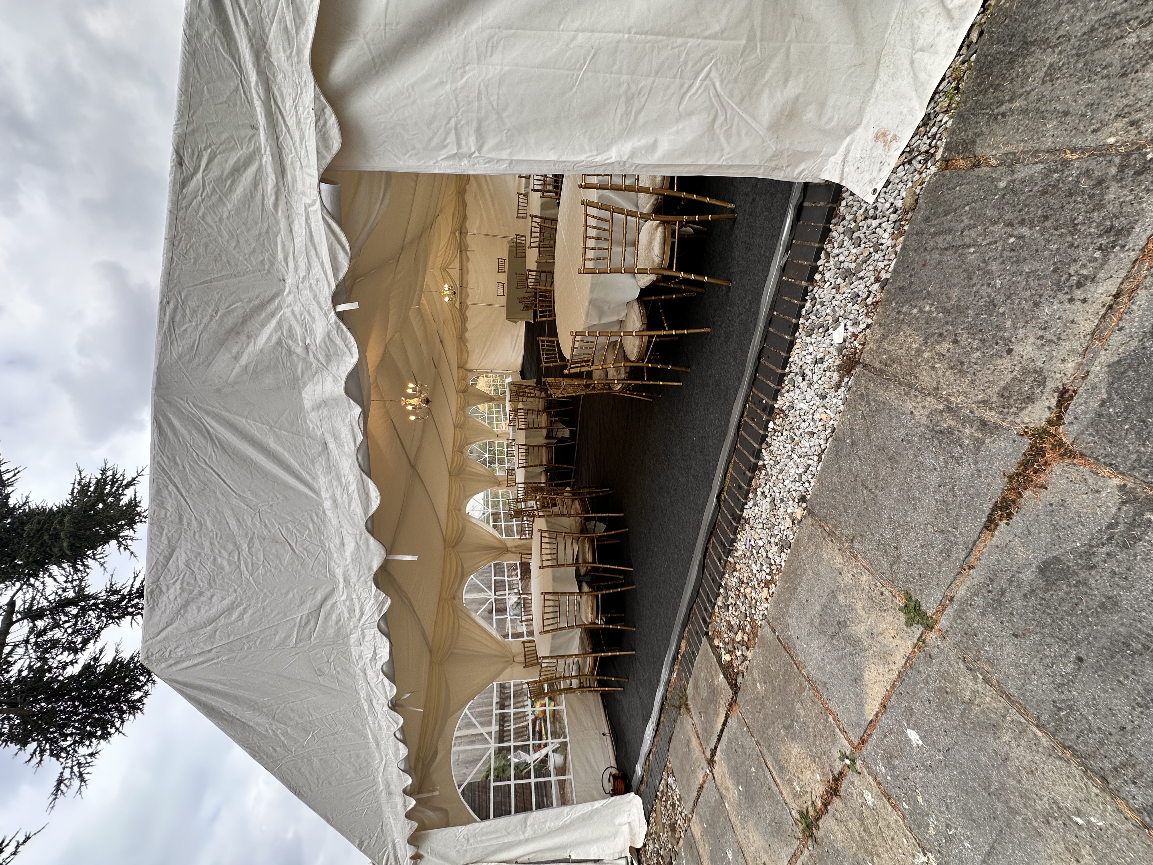 A spacious white tent features elegant round tables with crisp linen and gold Chiavari chairs, illuminated by soft chandelier lighting, perfect for catering events.
