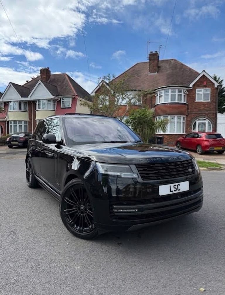 Black luxury Range Rover on a quiet suburban street, glossy black wheels and plate, framed by brick homes under bright daylight.