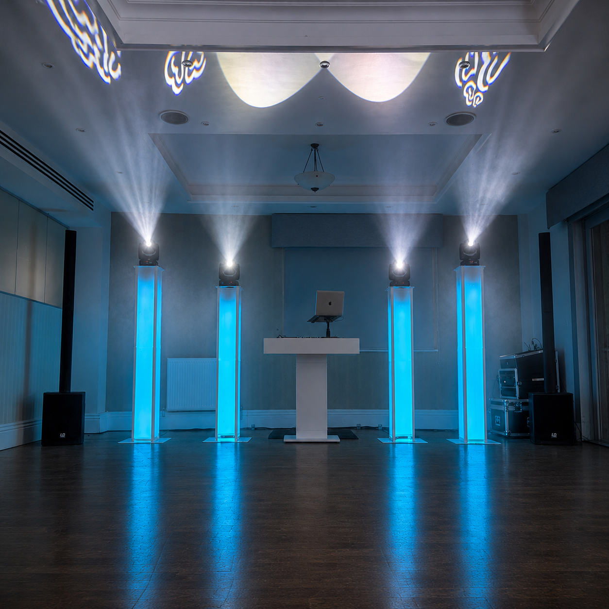 Modern DJ booth with glowing blue columns, centered laptop, and sweeping white spotlights in a sleek empty venue, ideal for event catering.