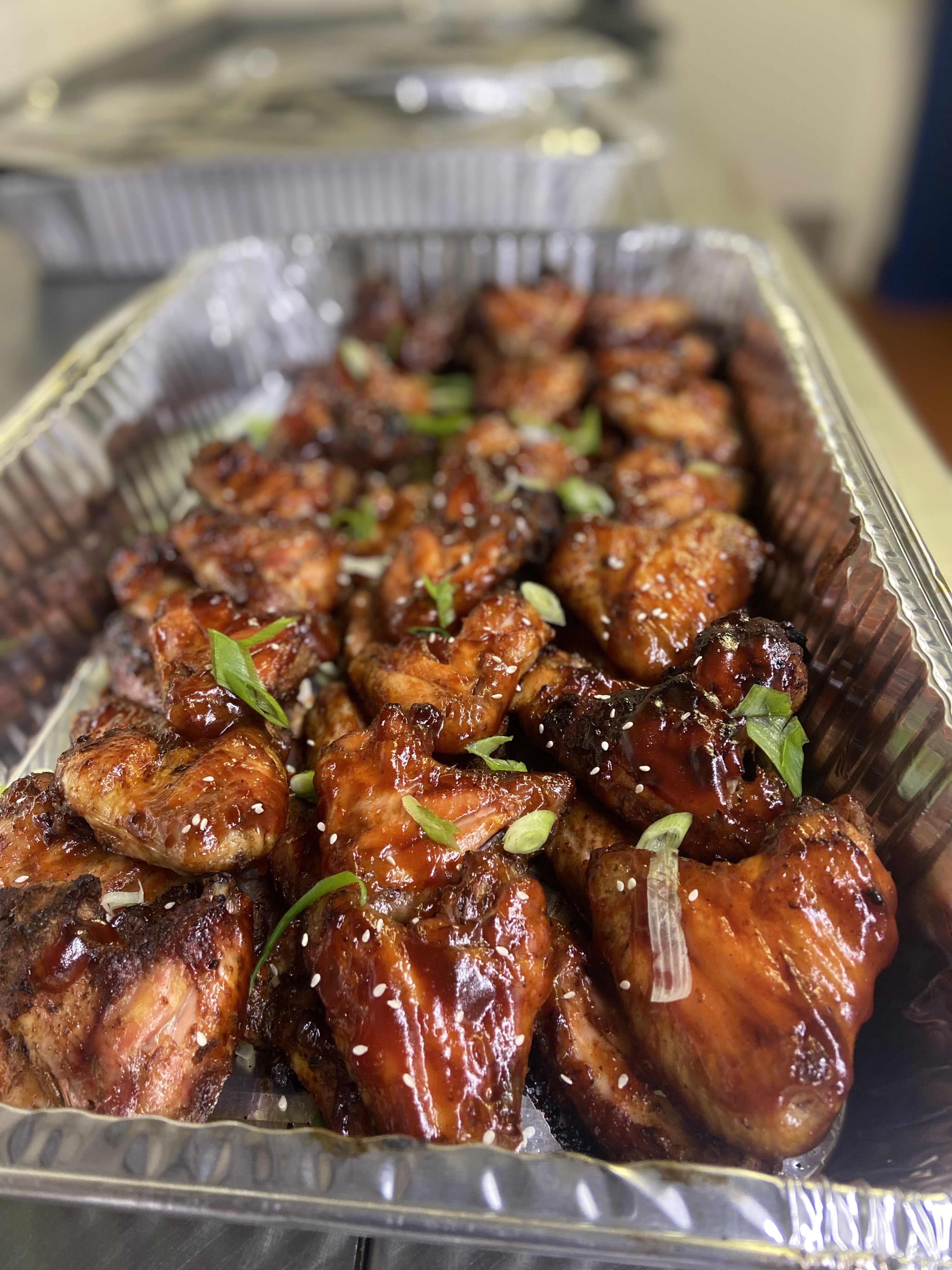 Succulent, glazed chicken wings are artfully arranged in a shiny aluminum tray, garnished with green onions and sesame seeds, showcasing quality catering.