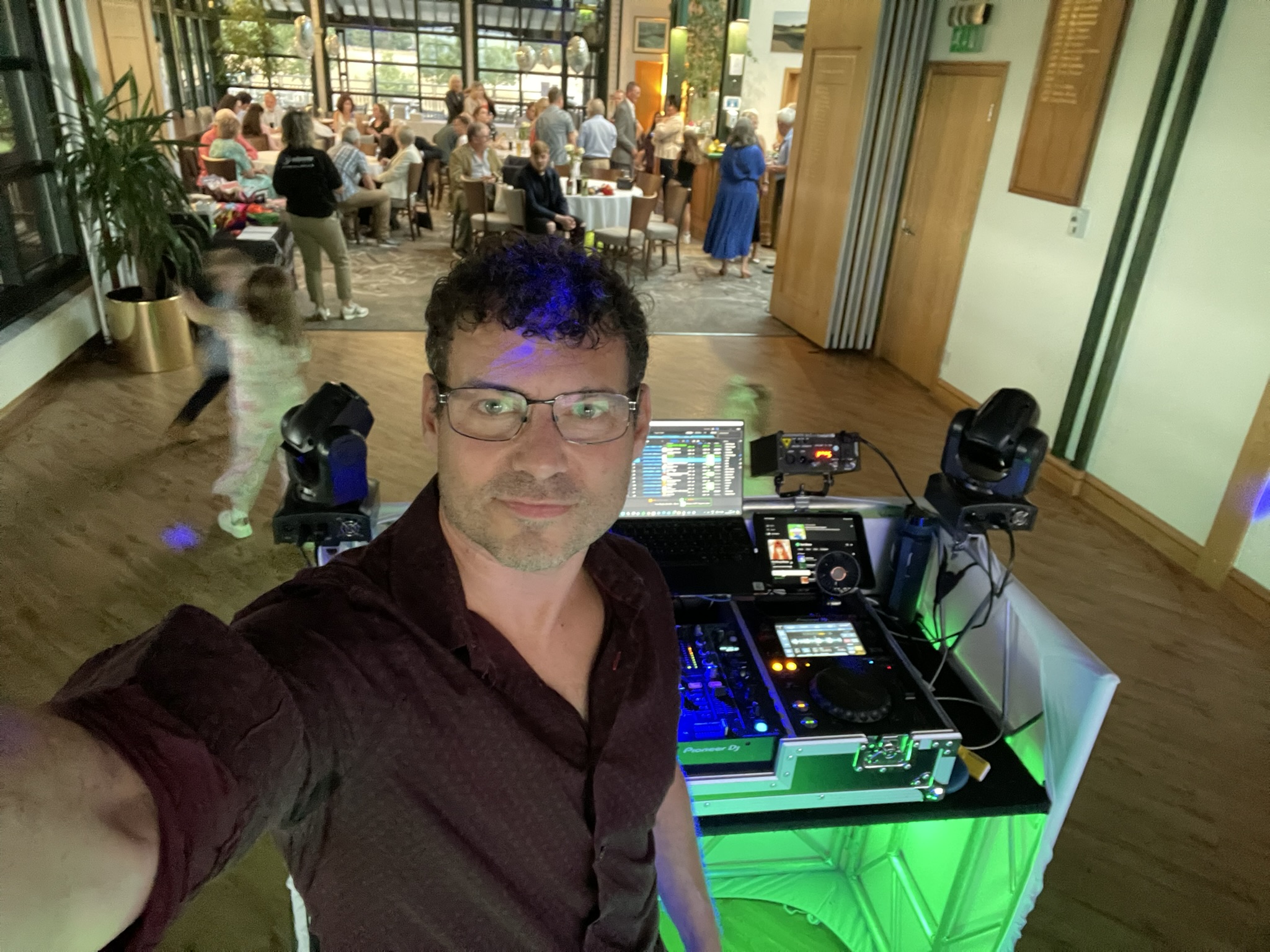 Selfie of a DJ behind a glowing Pioneer deck, laptop and moving lights, serving a lively catered reception with guests in a bright hall.
