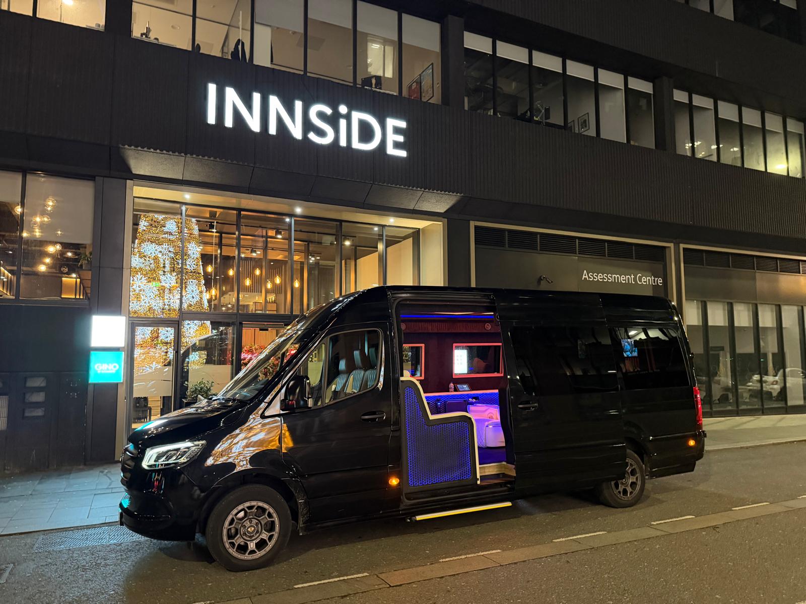 A sleek black van with vibrant blue lighting is parked outside a modern hotel featuring large glass windows, showcasing festive decor.
