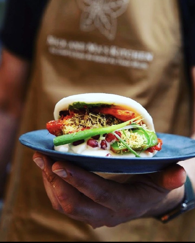 A hand holds a sleek blue plate featuring a bao filled with vibrant ingredients like green chili, crispy noodles, and bright pomegranate seeds, showcasing artisanal catering.