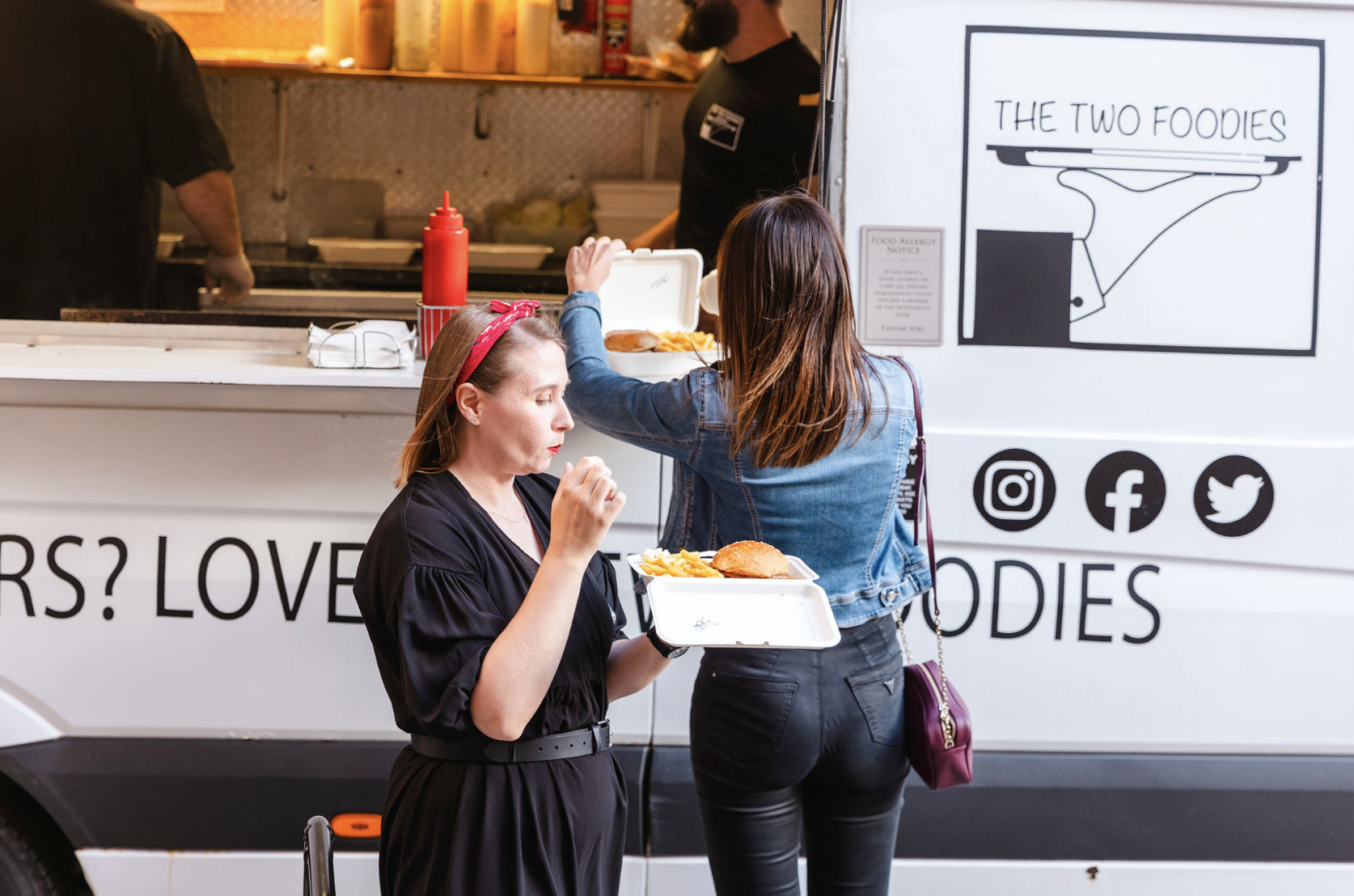 A food truck features sleek white panels, with two women enjoying fresh burgers and fries, vibrant red ketchup, under bright daylight, highlighting quality service.