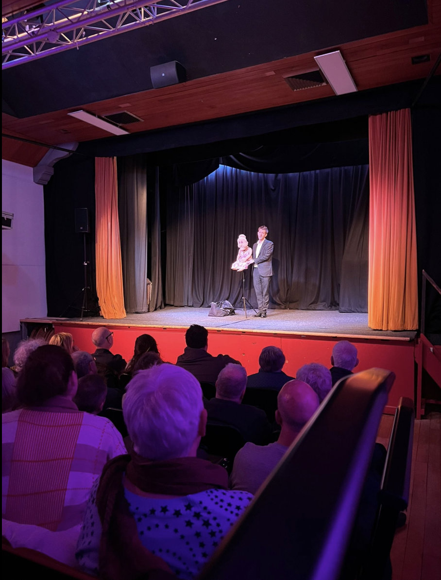 A vibrant theater features plush, curved seating in warm tones, illuminated by soft blue lights, as a performer joyfully holds a puppet.