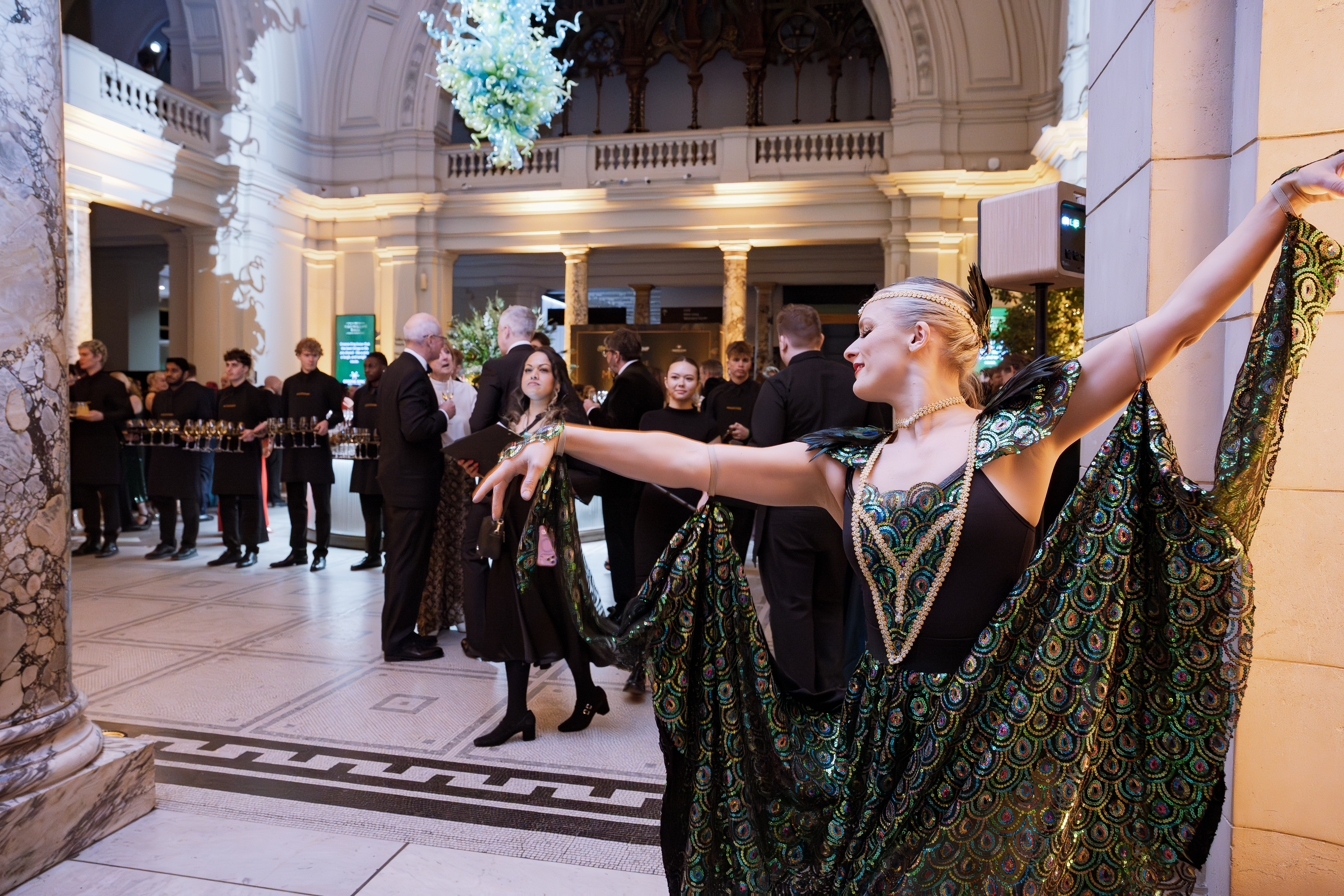 A dazzling performer in a sequined green costume captures attention amidst guests enjoying cocktails, with elegant decor and warm lighting.