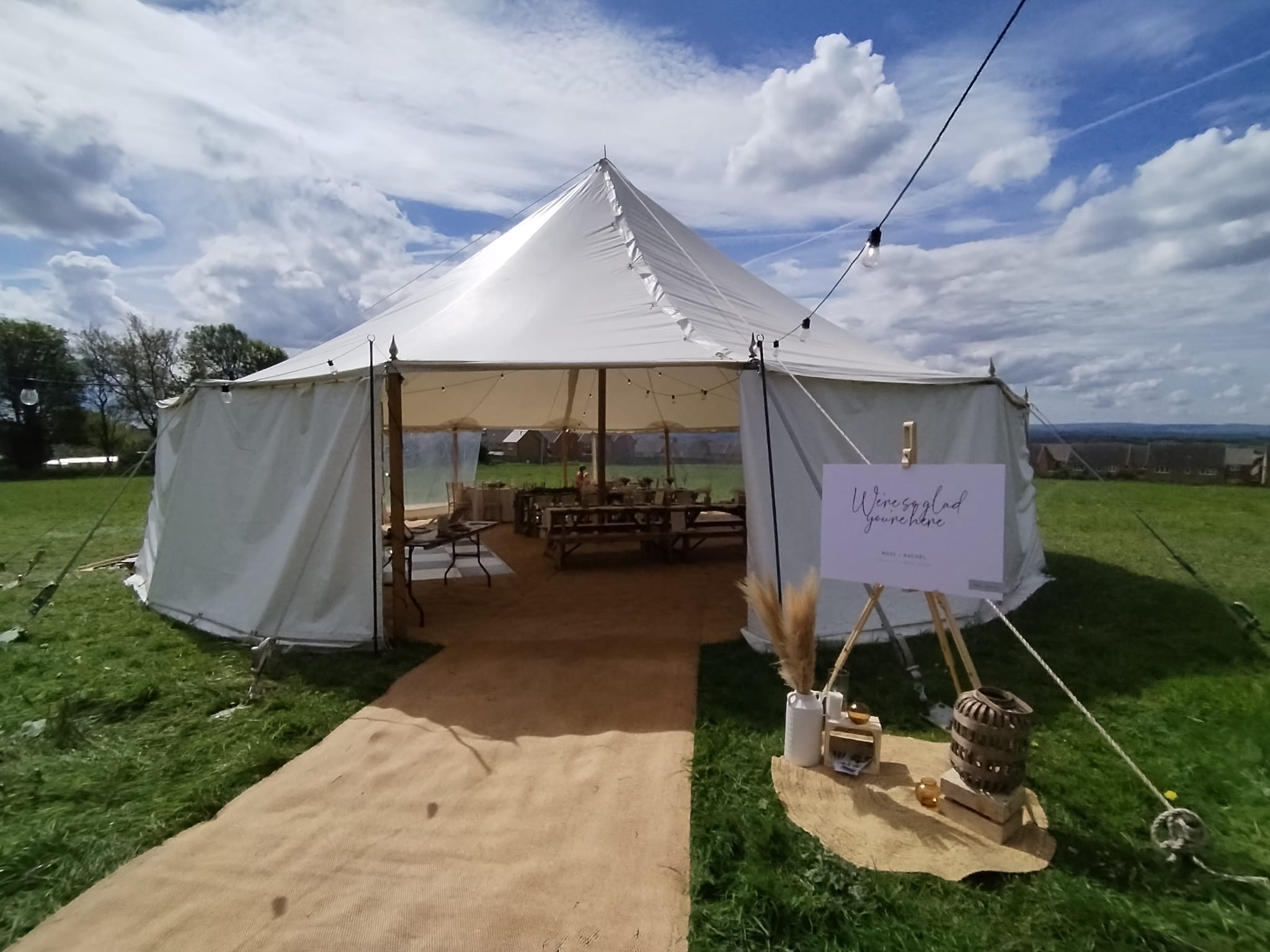 A spacious white marquee tent sits on lush green grass, surrounded by trees under a bright, partly cloudy sky, perfect for outdoor catering.
